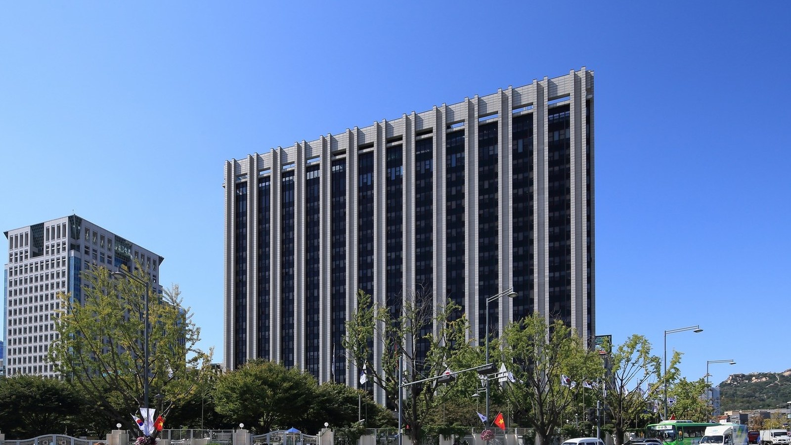 The main building of Government Complex Seoul photographed from ground level against a clear blue sky. The tower's facade is divided into narrow vertical concrete fins running its full height, with bands of dark glass recessed between them, and the fins step out above the roofline. A lower pale stone office block stands to its left. Ginkgo trees, street lights and a row of small flags on poles run along the road in front of it, a green city bus and several vans pass at the bottom right, and low wooded hills close the horizon on the right.