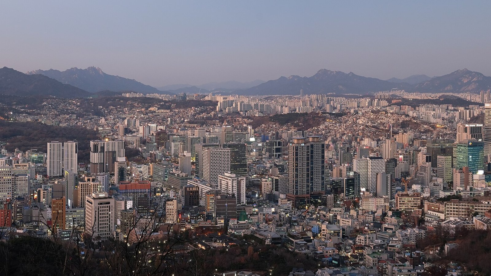 A view over central Seoul at dusk from Namsan, dense mid-rise blocks and office towers filling the valley below and a hazy mountain ridge along the horizon.