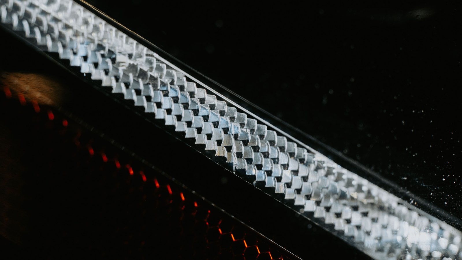 A close view of a clean machined metal band with a repeating chevron pattern catching bright light against a dark background, with an out of focus red reflector strip at the lower left.