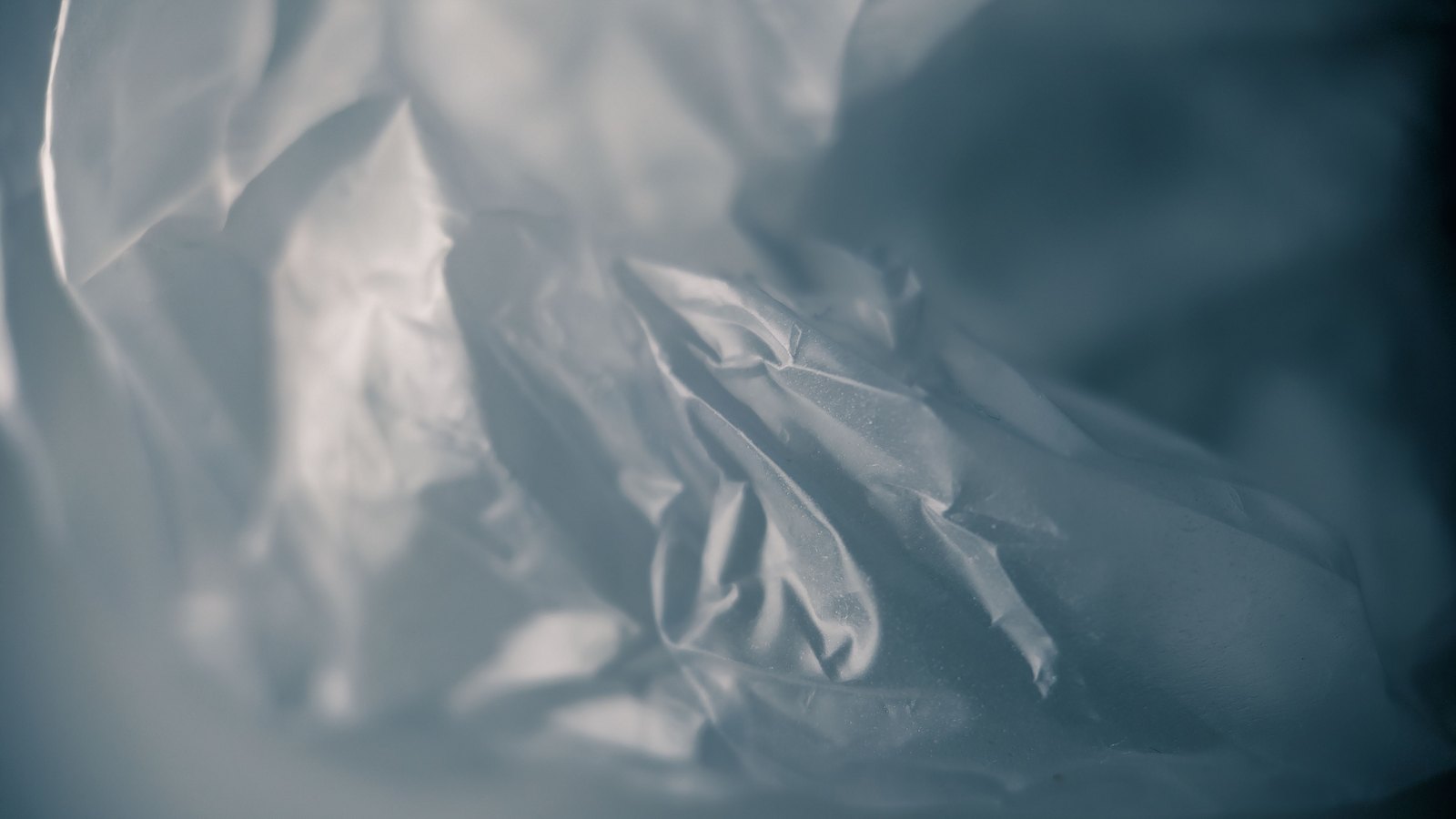 A macro view of crumpled translucent plastic film in cool blue-grey, its folds catching soft light and falling out of focus toward the edges.
