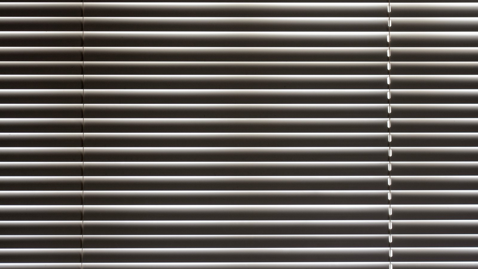 A close photograph of a fully closed venetian blind filling the frame, its pale horizontal slats overlapping so that nothing behind them is visible, with a thin vertical line of daylight showing through the cord holes towards the right edge.