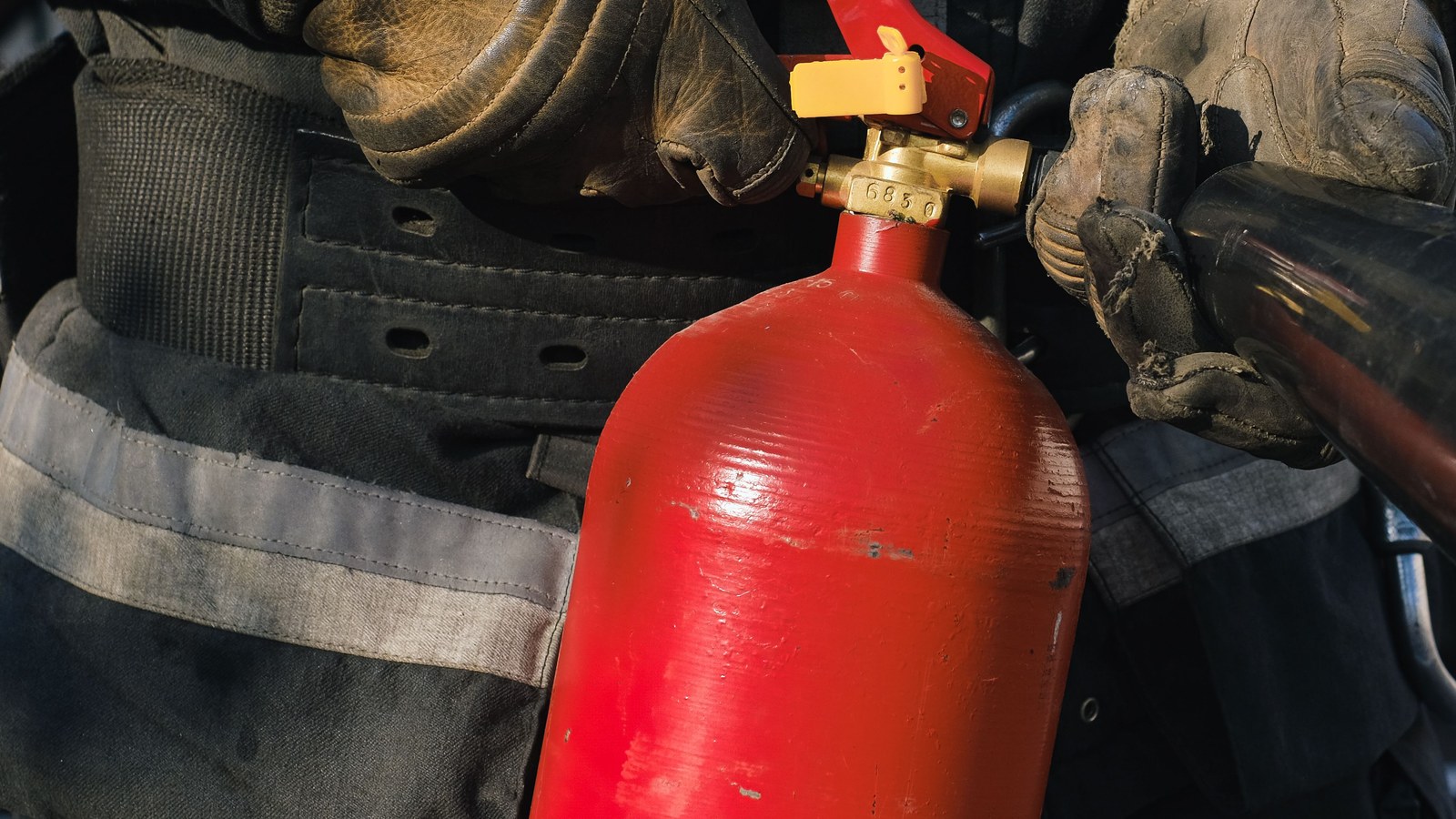 Gloved hands holding the valve and horn of a red fire extinguisher.