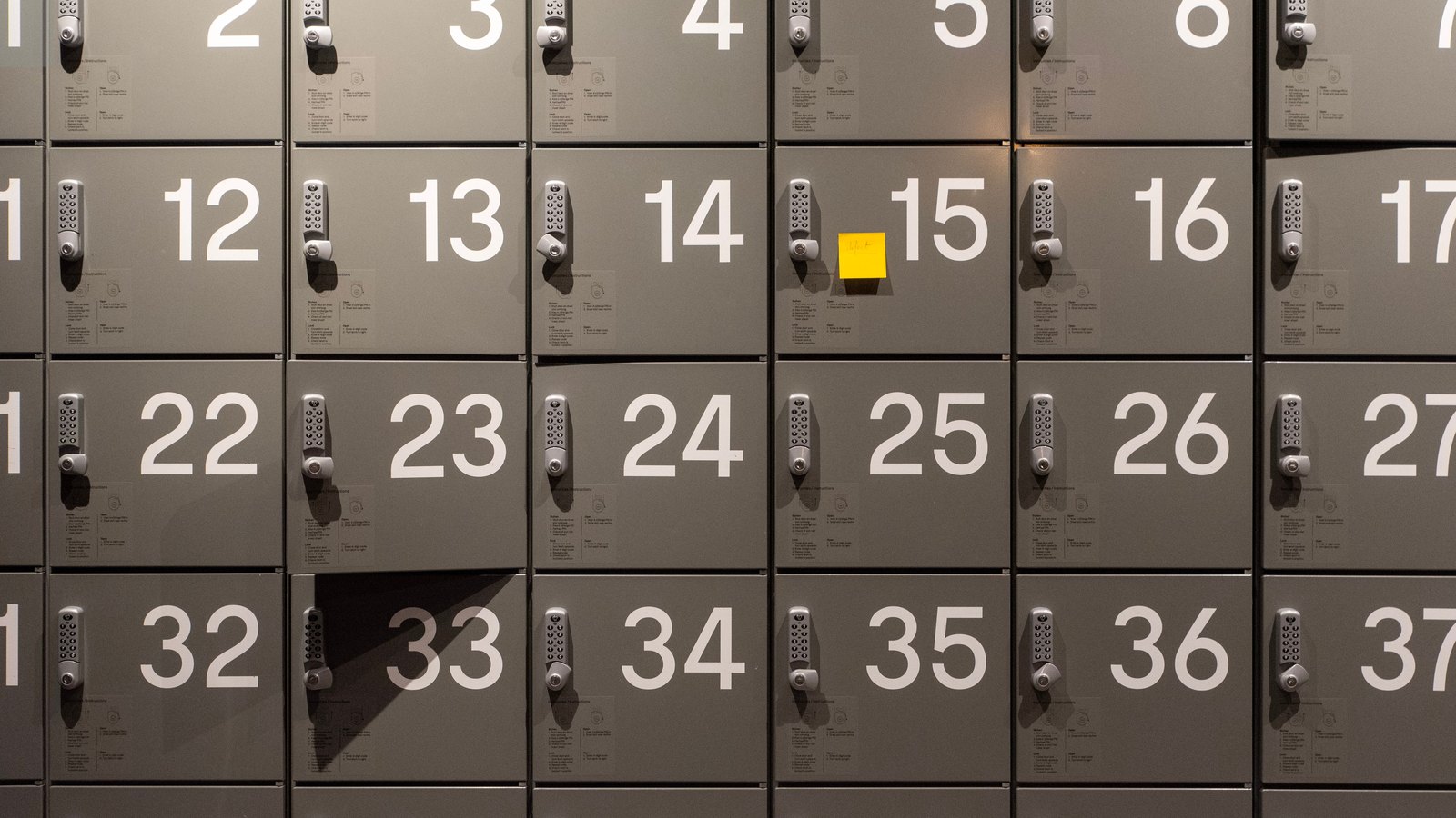A grid of grey metal locker doors, each carrying a large white number and a small silver keypad combination lock with a printed instruction label beneath it. One door stands ajar and a blank yellow sticky note is stuck beside the lock on another.