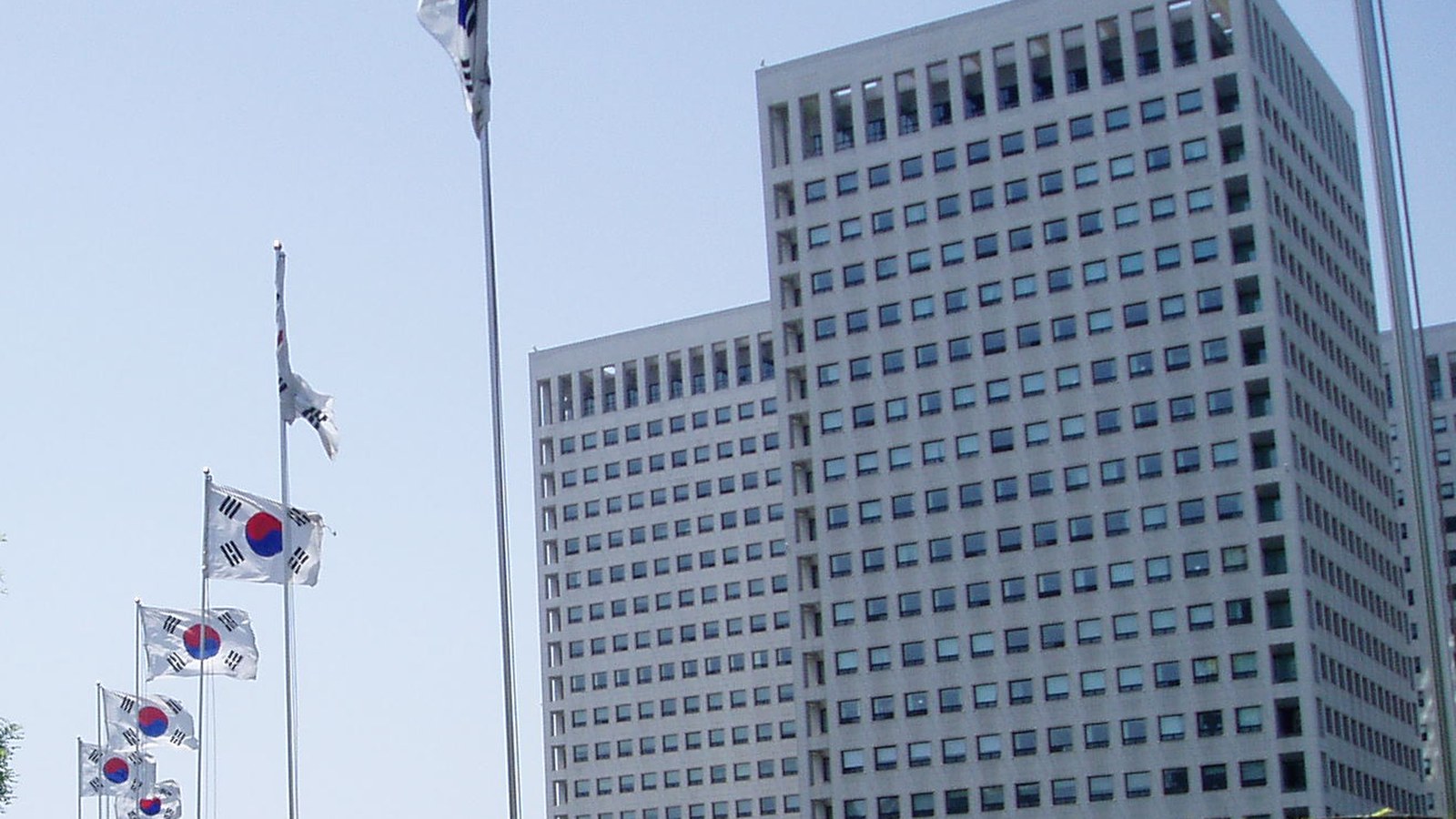 Two pale high-rise towers of the Government Complex Daejeon beside a row of South Korean flags flying on tall poles under a clear sky.