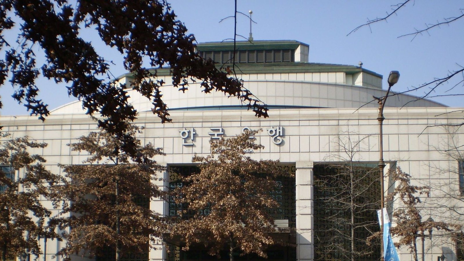 The Bank of Korea's Daegu office, a pale stone building with a shallow domed roof and the bank's Korean name above the entrance, framed by autumn trees.