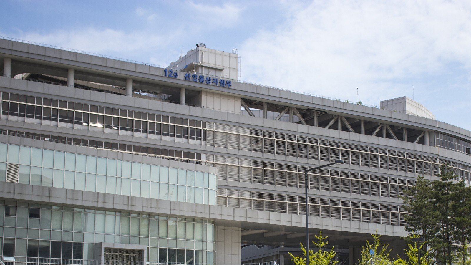 A long curving grey government building at the Sejong complex with horizontal louvres and glass bands, its roofline sign reading Building 12 and the trade ministry's earlier Korean name, under a lightly clouded sky.