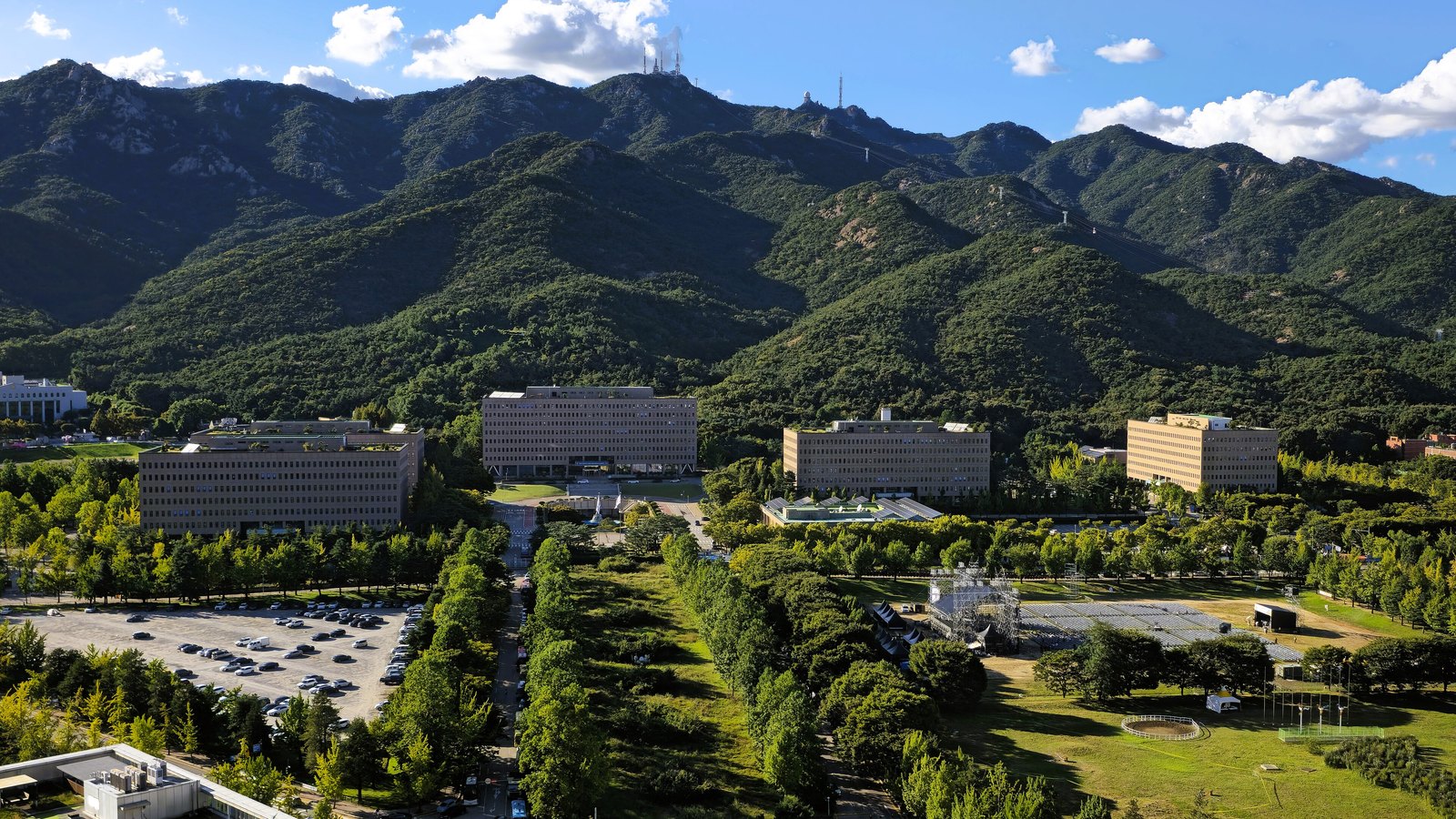 An elevated view of four long government office blocks in pale stone standing among trees and lawns at the foot of a forested mountain ridge, with a car park and an avenue of trees in the foreground and scattered cloud over the summits. No building carries a legible name.