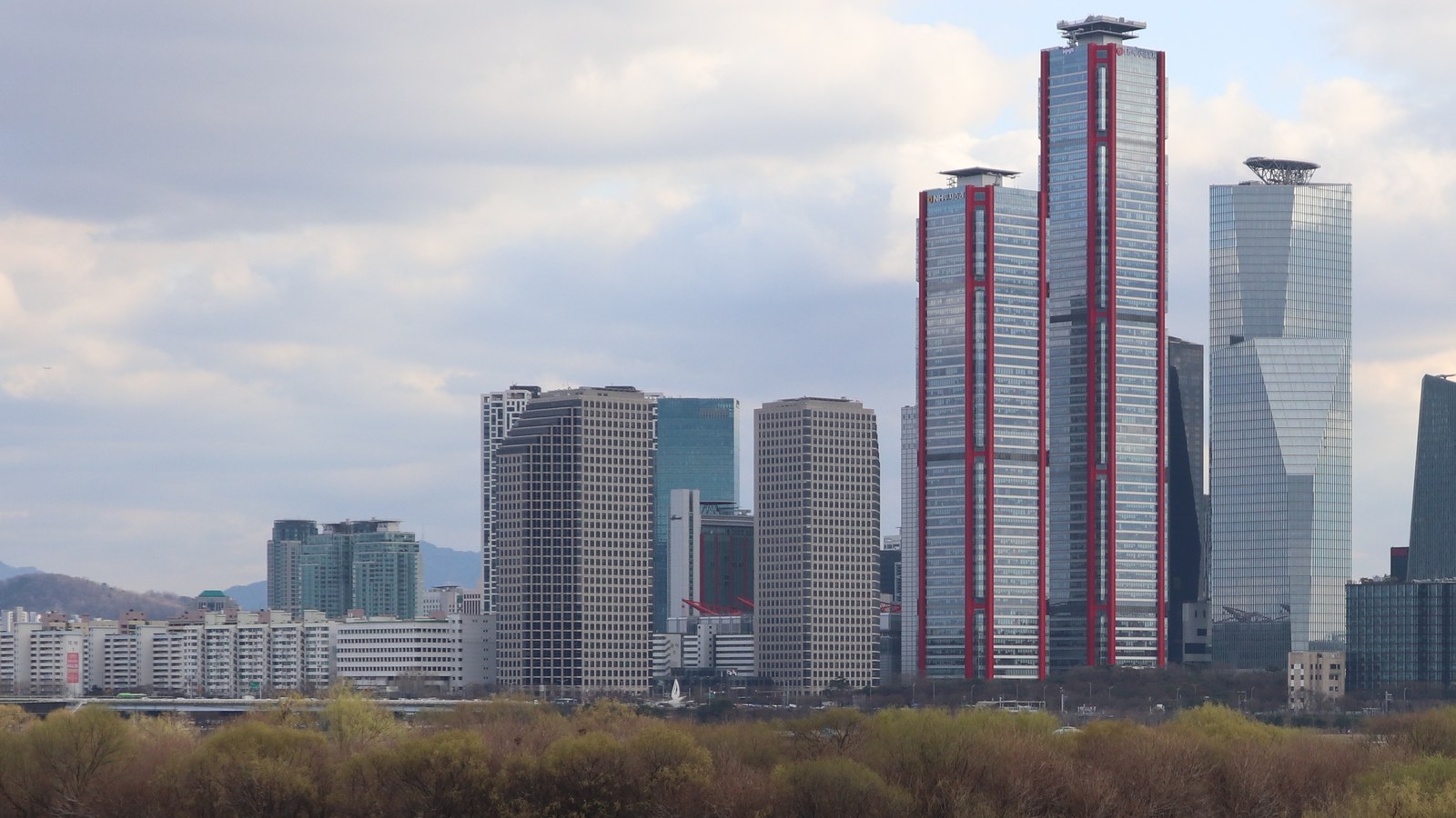 The Yeouido financial district of Seoul photographed across the Han River on a clear spring day. Willow scrub and a sandbank fill the near bank, with a line of white apartment blocks and pale grey office slabs behind them, and towards the right the two red framed towers of the Parc1 complex and a faceted glass tower standing above everything around them. Small rooftop company marks sit near the tops of the tallest towers and are not readable at this size. Low hills close the horizon on the left.