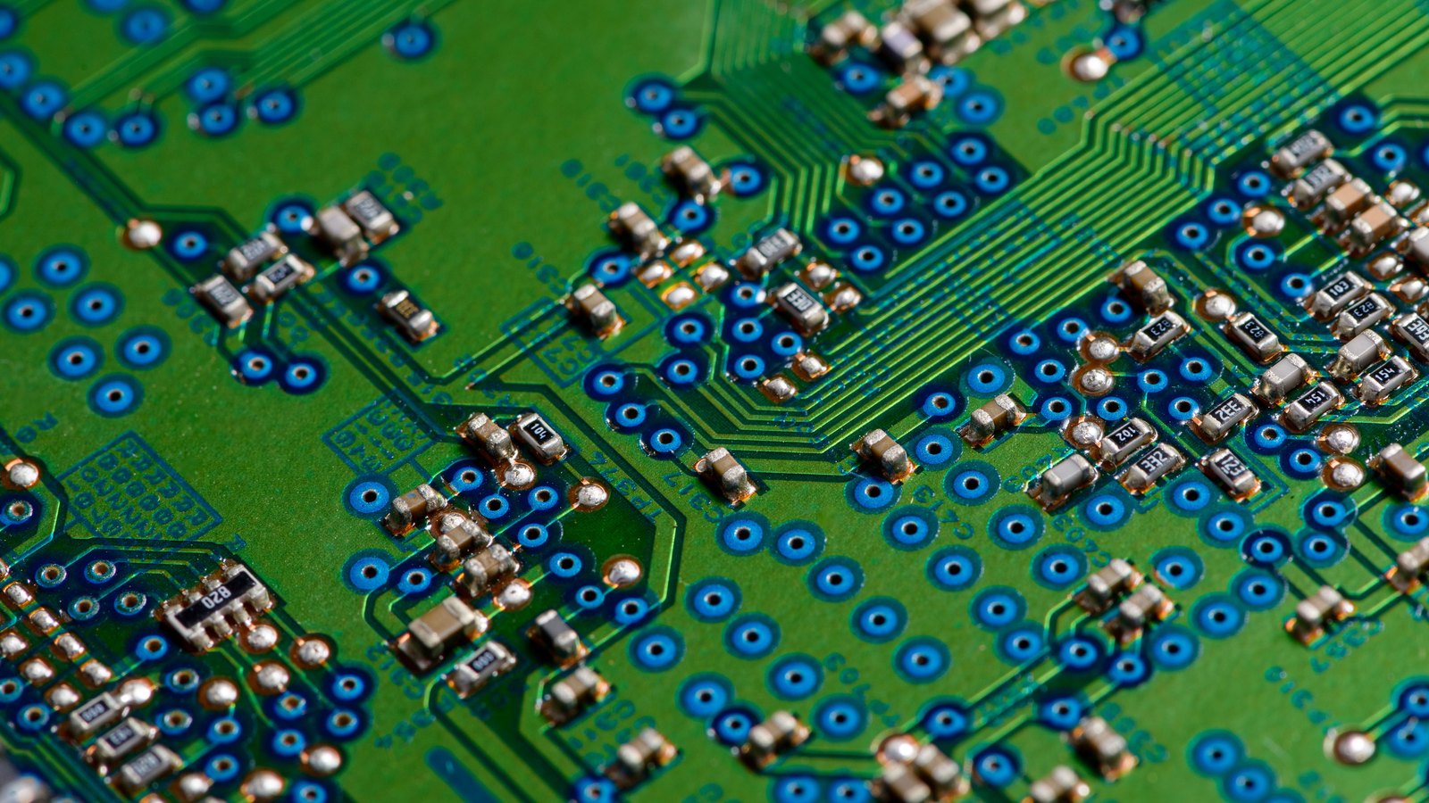 A close macro of a green printed circuit board lit from above, showing rows of blue ring-shaped solder pads, fine copper tracks fanning out across the board, and dozens of small soldered surface-mount components marked with numeric value codes.