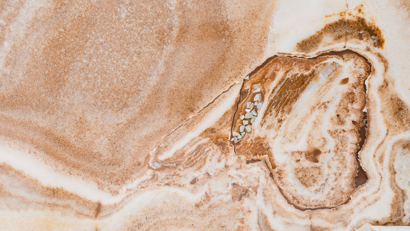 A close photograph of a polished onyx marble surface, its pale cream ground crossed by broad caramel and rust coloured bands that curl in tight rings around a darker oval formation to the right of the frame.