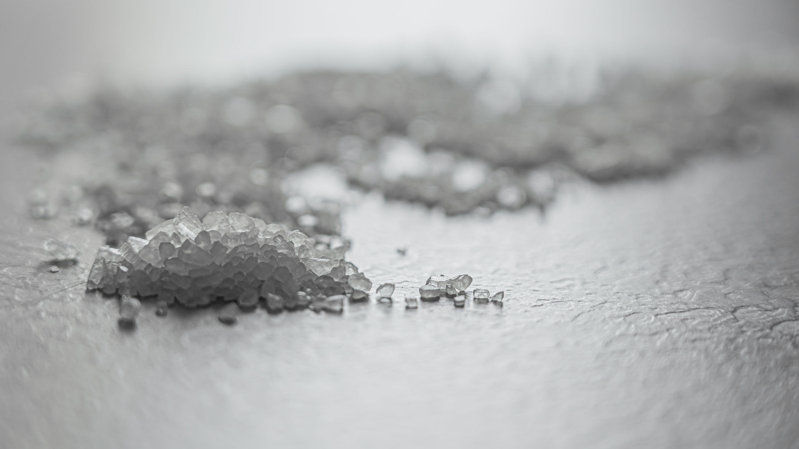 A close monochrome photograph of coarse salt crystals on a pale grey surface, a small heap of them sharp in the left foreground with single grains trailing away to the right and the rest of the scatter falling out of focus behind.