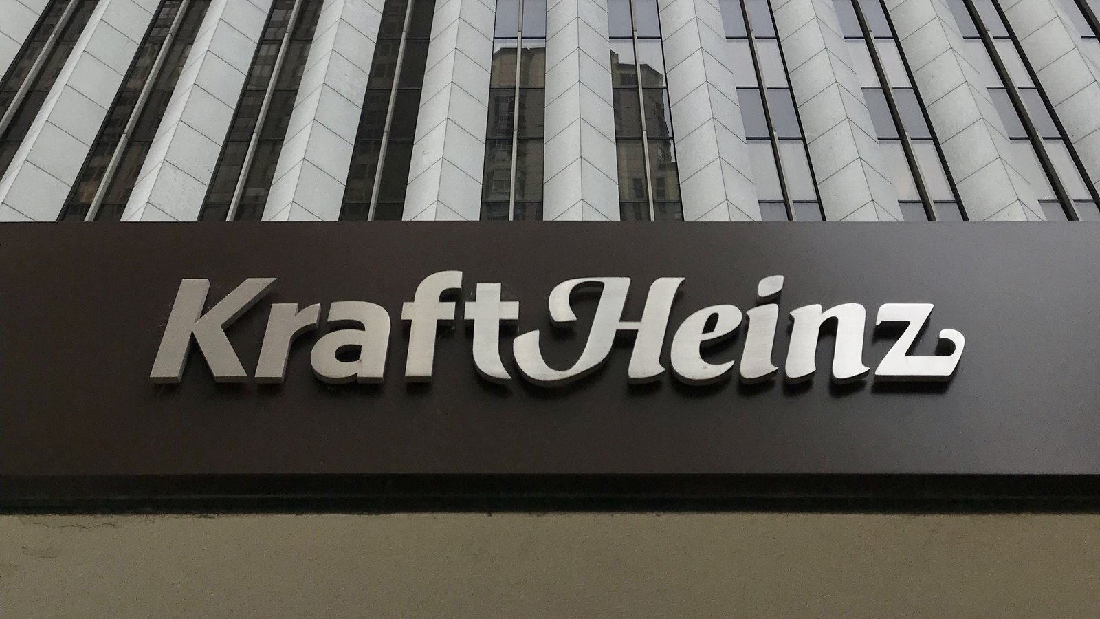 The Kraft Heinz wordmark in brushed steel letters mounted on a dark brown entrance canopy, with the pale ribbed facade of the tower above it rising out of the top of the frame, photographed from street level looking up.