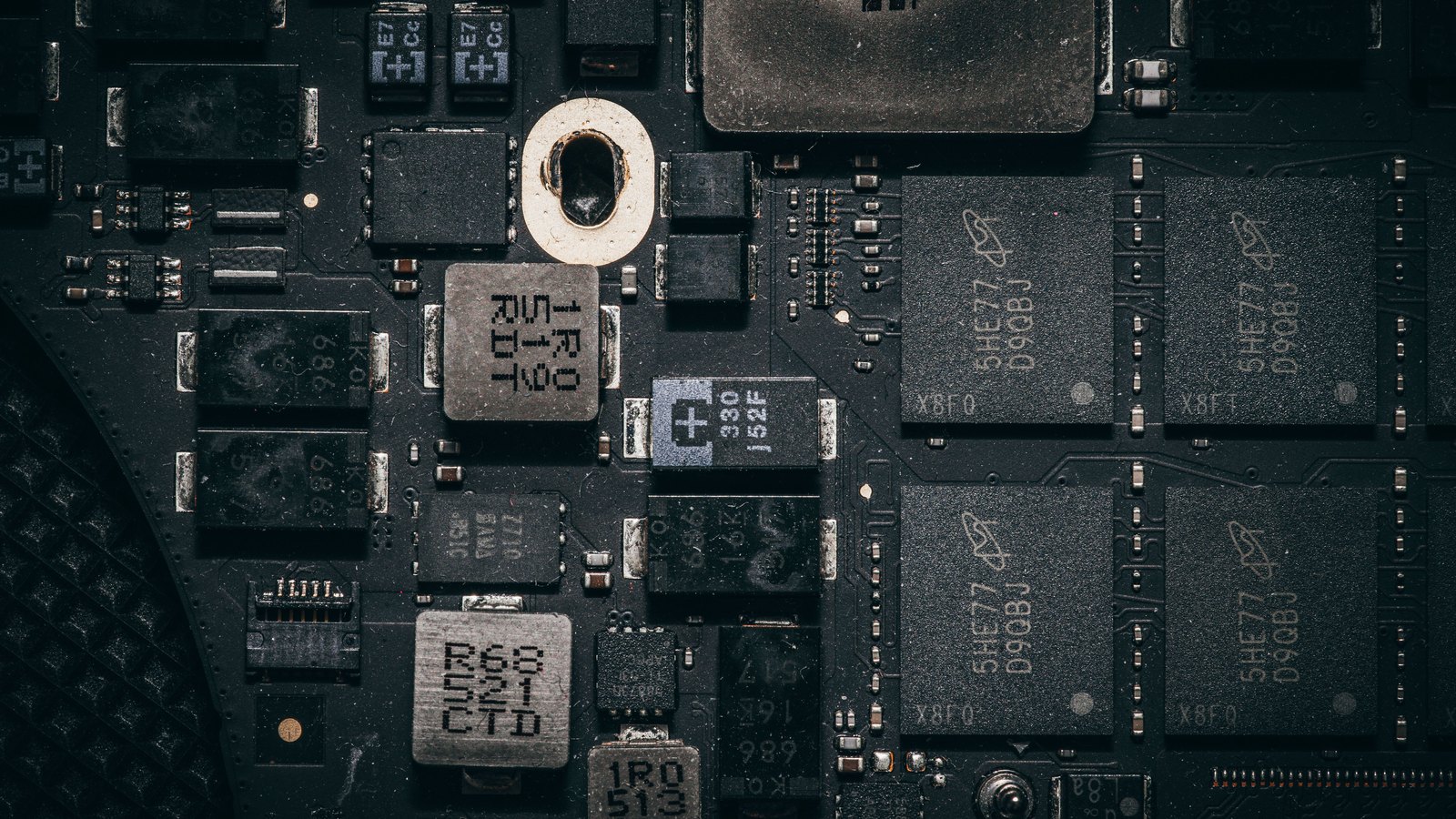 A tight overhead macro of a dark circuit board, with four large black memory packages ranked in a grid on the right and an array of smaller capacitors, inductors and diodes across the left, all carrying small printed part codes.