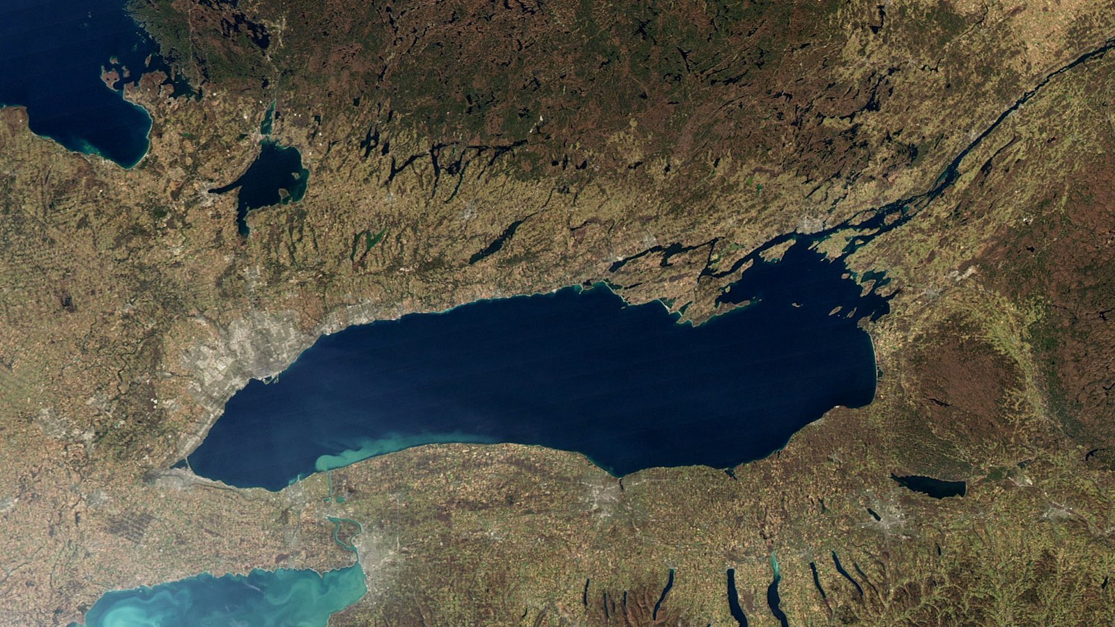 A satellite view of Lake Ontario in full, the lake a deep navy oval running west to east across the frame, with the Niagara peninsula and the eastern end of Lake Erie at lower left, the Finger Lakes as thin dark slivers at lower right, and the outlet to the St. Lawrence at upper right. Sediment shows pale turquoise along the southwestern shore.