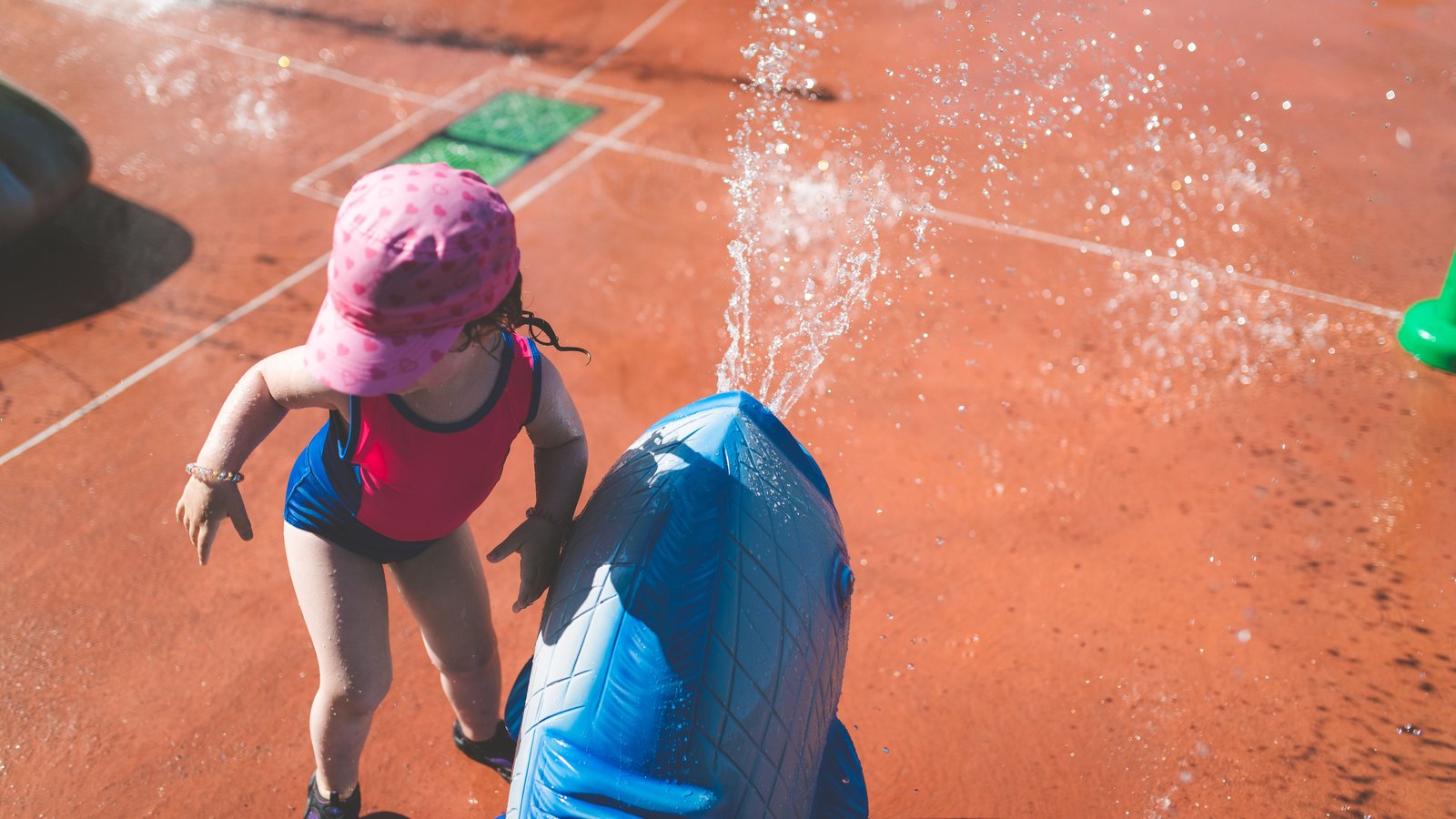 Seen from above, a small child in a pink sun hat and a red and blue swimsuit stands on a sun bright orange splash pad beside a blue whale shaped water toy that is shooting a jet of water into the air. The child is turned away from the camera and no face is visible. No signage, lettering, logo, street, building or landmark of any kind appears in the frame. Stock photograph, not a photograph of this event.