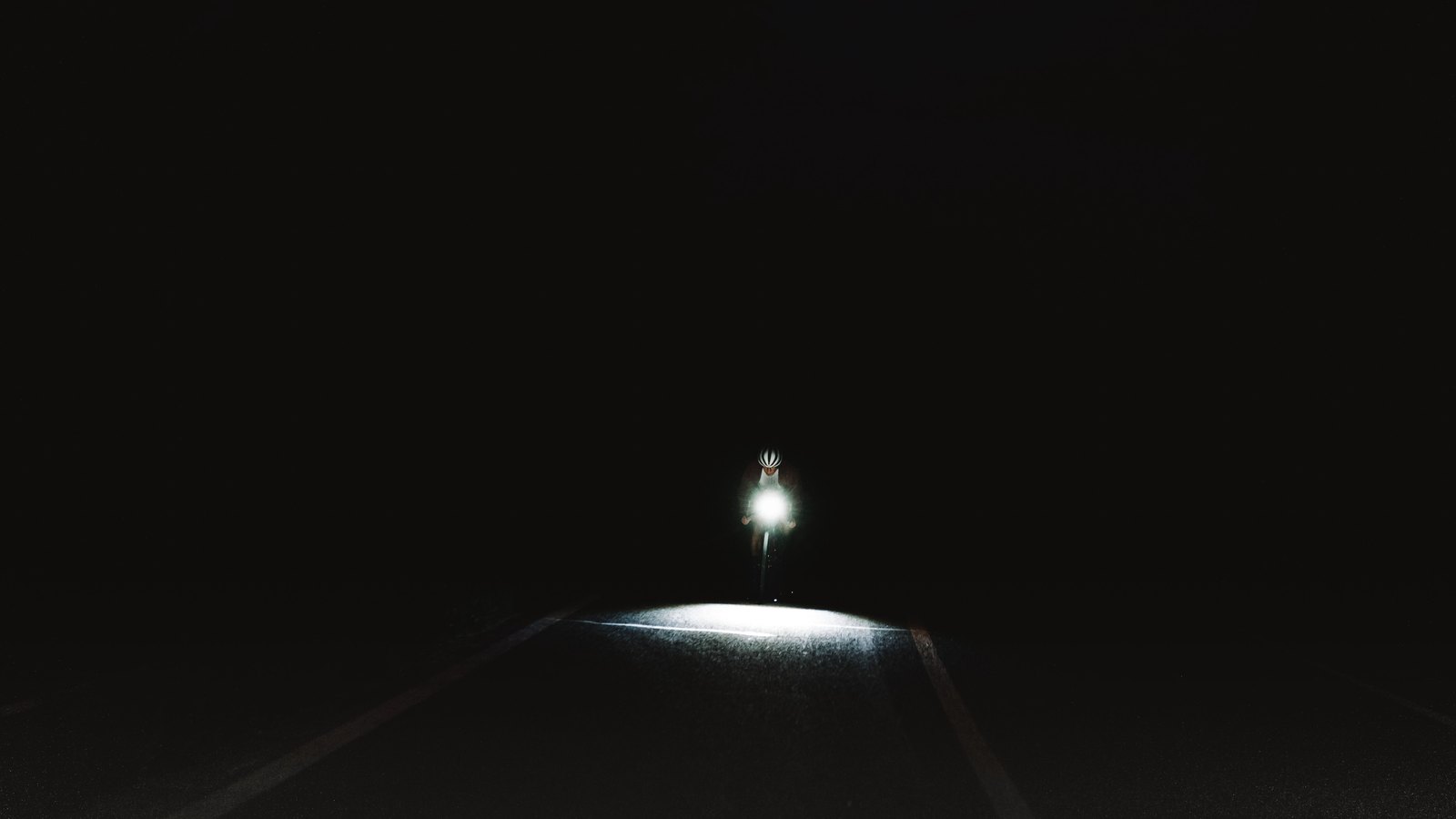 A single bicycle headlight on an unlit road at night, the rider not visible beyond the glare. Stock photo, not a photograph of this event.