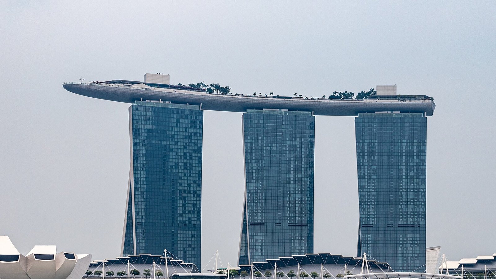 The three glass hotel towers of Marina Bay Sands in Singapore joined at the top by a long cantilevered sky deck lined with palm trees, under a pale grey sky, with the white petal roof of the ArtScience Museum at lower left.