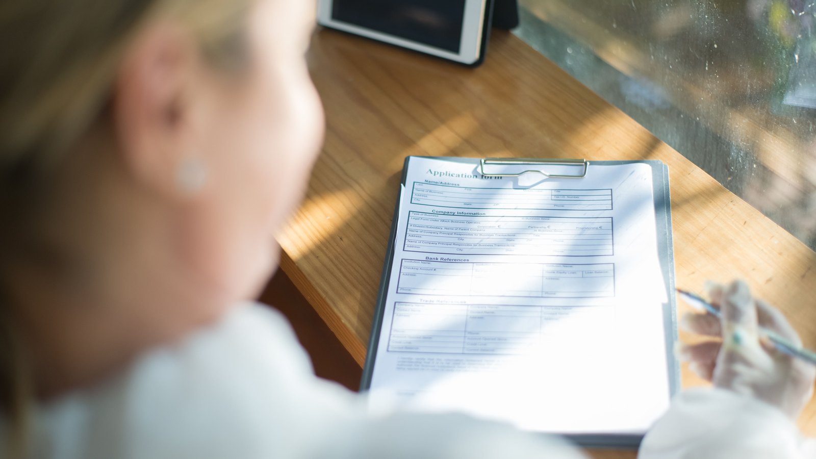 A person sits at a wooden table beside a bright window, seen from behind and over the shoulder so that only blurred hair and the edge of a cheek are visible, holding a pencil above a printed application form clipped to a board. Sunlight falls across the paper and a closed tablet stands on the table behind it. Stock photo, not a photograph of this event.