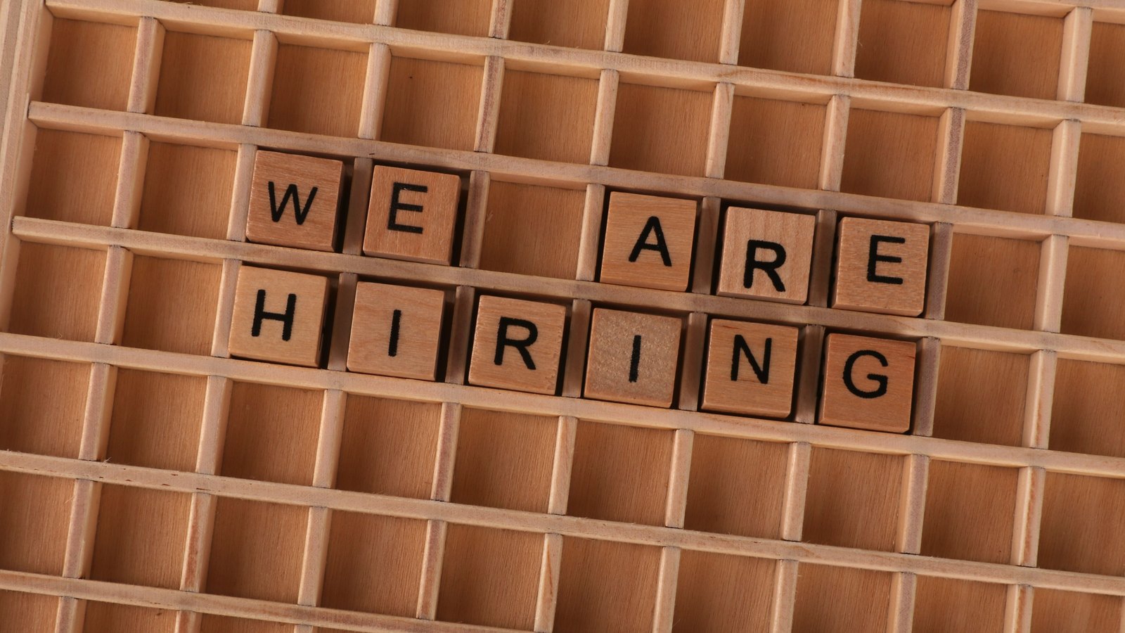 Wooden letter tiles on a grid board spelling We Are Hiring, not a photograph of this event.