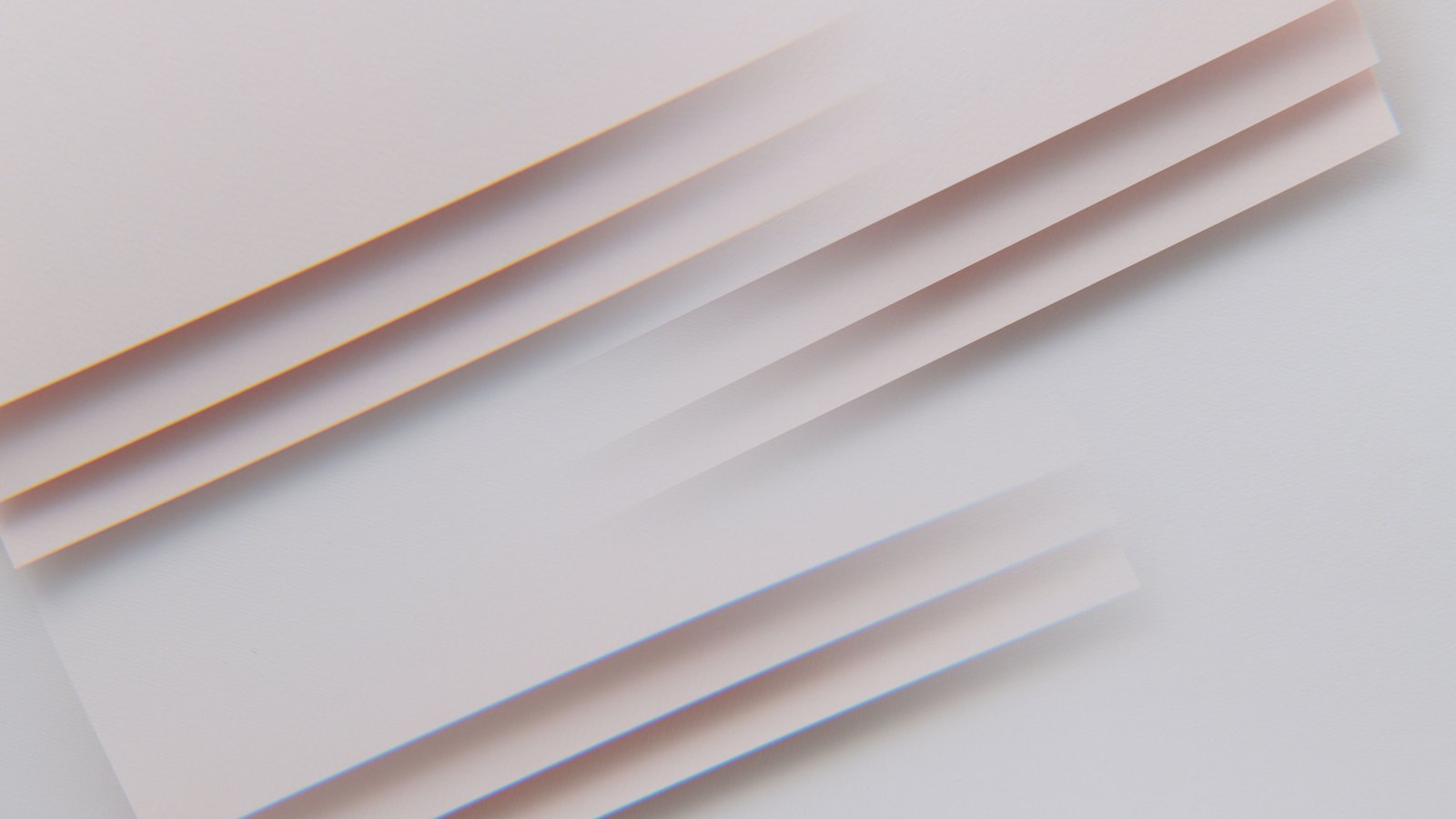 Several plain sheets of pale paper lying fanned across one another on a light grey surface, photographed close and at an angle so only their overlapping edges show. No writing, markings, people or objects are in view.