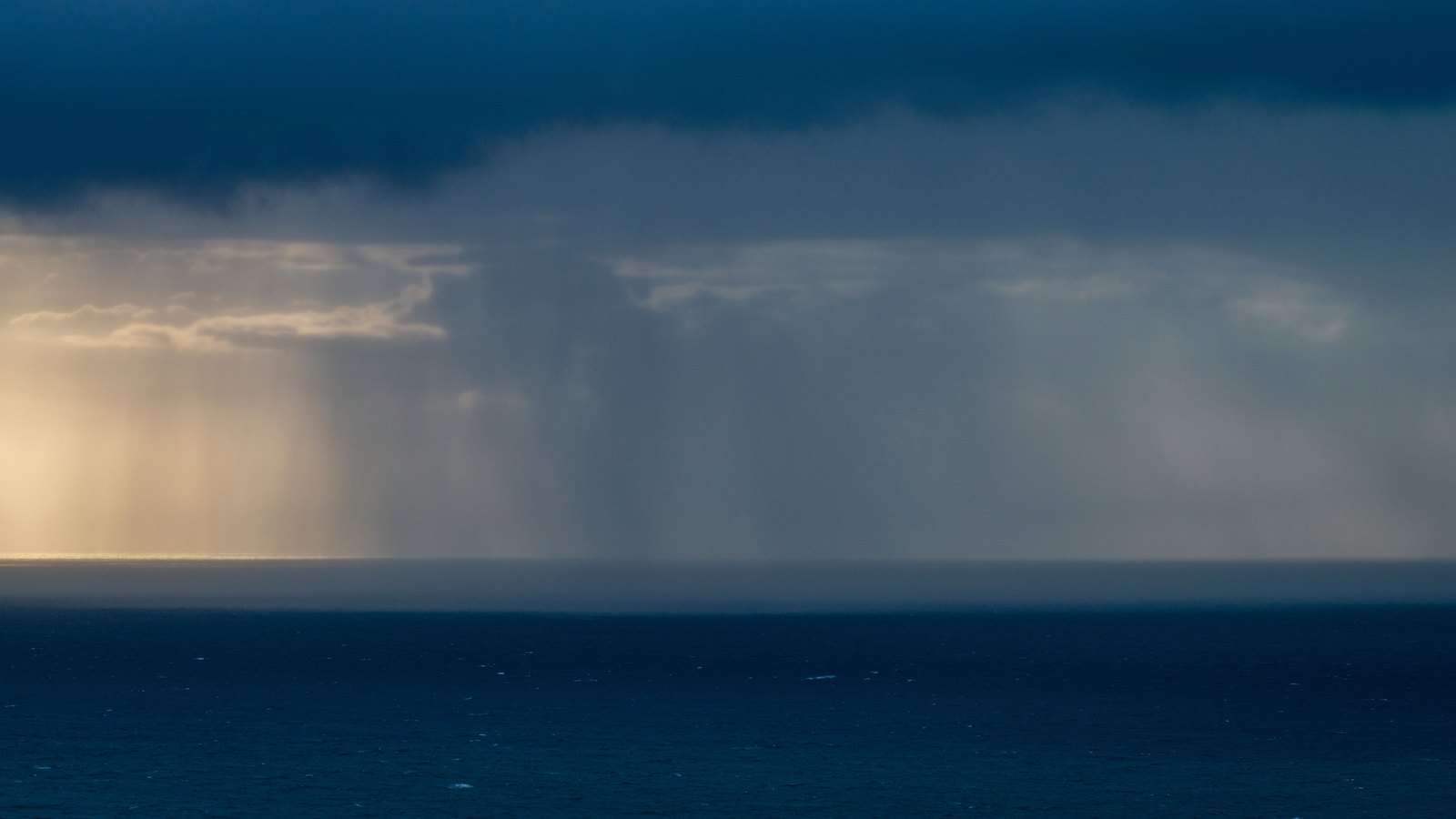 A dark cloud base sits low over open water with a broad shaft of rain falling from it to the horizon, sunlight breaking through at the left edge and deep blue sea in the foreground, with no people, vessels, buildings, signs or text in view.