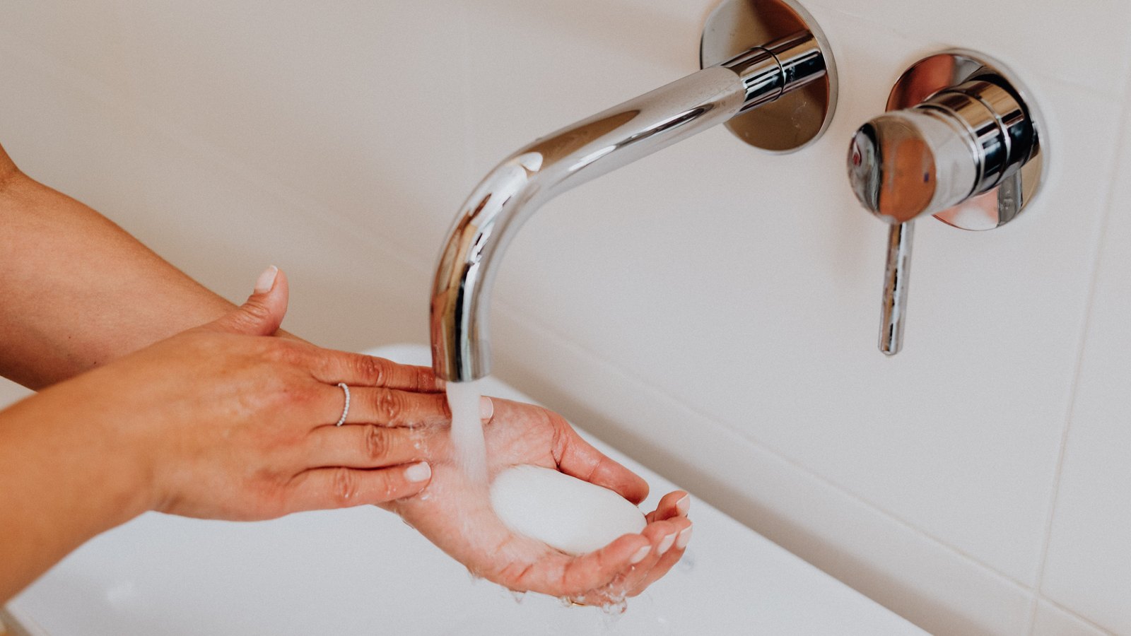 A pair of hands held under a wall mounted chrome tap in a white tiled bathroom, one hand cupping a white bar of soap while running water falls across both. No faces, signs or text are in view. A generic illustration, not a photograph taken in Lee or Collier County.