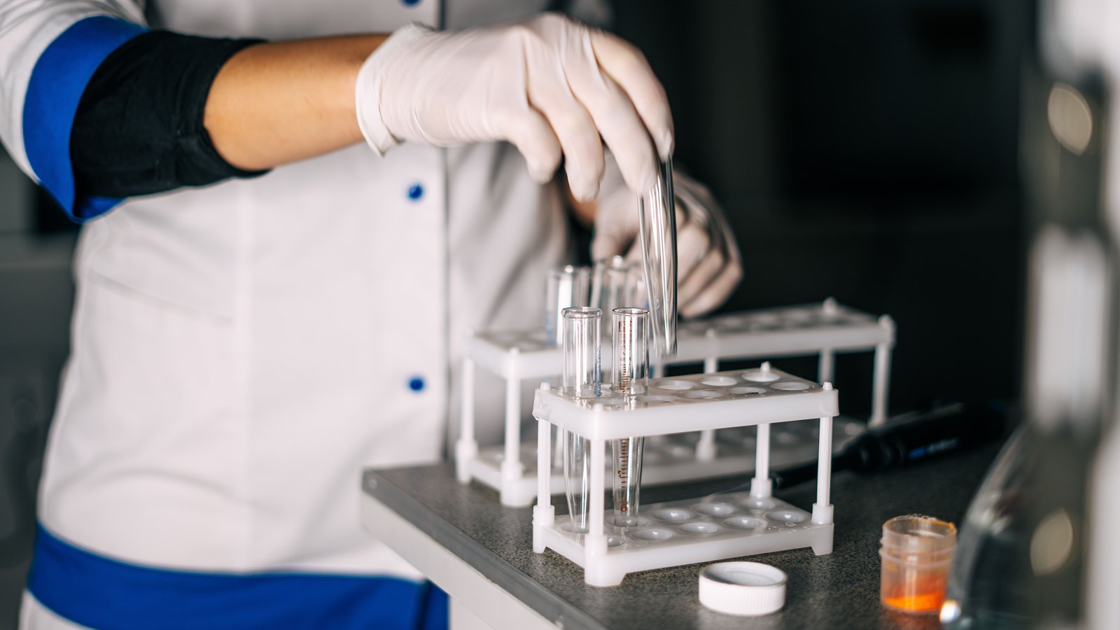 A gloved hand in a white lab coat with blue trim lowering a pair of empty glass test tubes into a white plastic rack on a dark laboratory bench, with a small amber specimen jar and its white cap standing beside the rack. No face, signage, logo or location is in view. A generic illustration of laboratory specimen testing, not a photograph of this event.