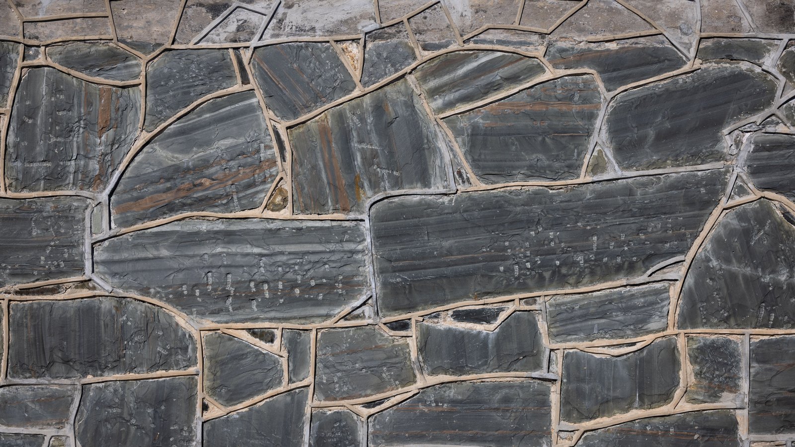 A flat wall of irregular grey slate flagstones filling the whole frame, each slab cut to a different angular shape and separated by pale sand coloured mortar joints, with faint rust and ochre banding running through some of the stone. No text, signage, lettering, person, vehicle, building or identifiable place is visible. A plain masonry surface used as illustration, not a photograph of any location, person or event in this case.