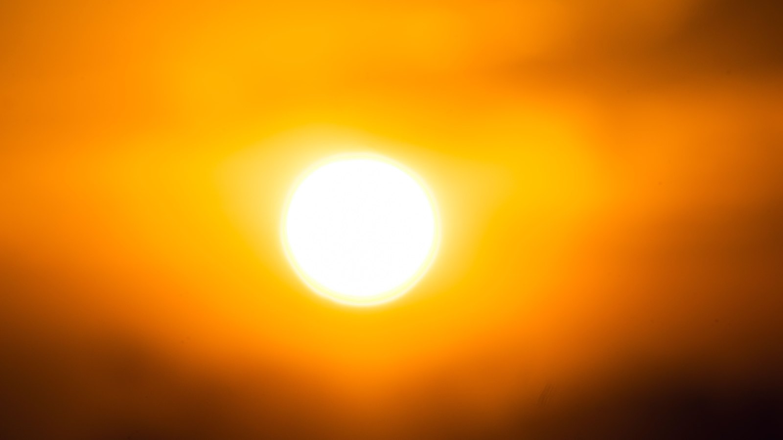 A blown-out white disc of sun sitting slightly left of centre in a sky of deep saturated orange, the glare fading outward through yellow into darker orange and near-brown along the top and bottom edges. Faint horizontal bands of haze cross the frame on either side of the sun. No ground, horizon, buildings, people, signage or text are in view.