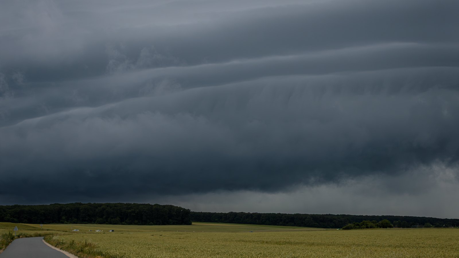 A dark shelf cloud over a flat field and narrow road, not a photograph of this event.