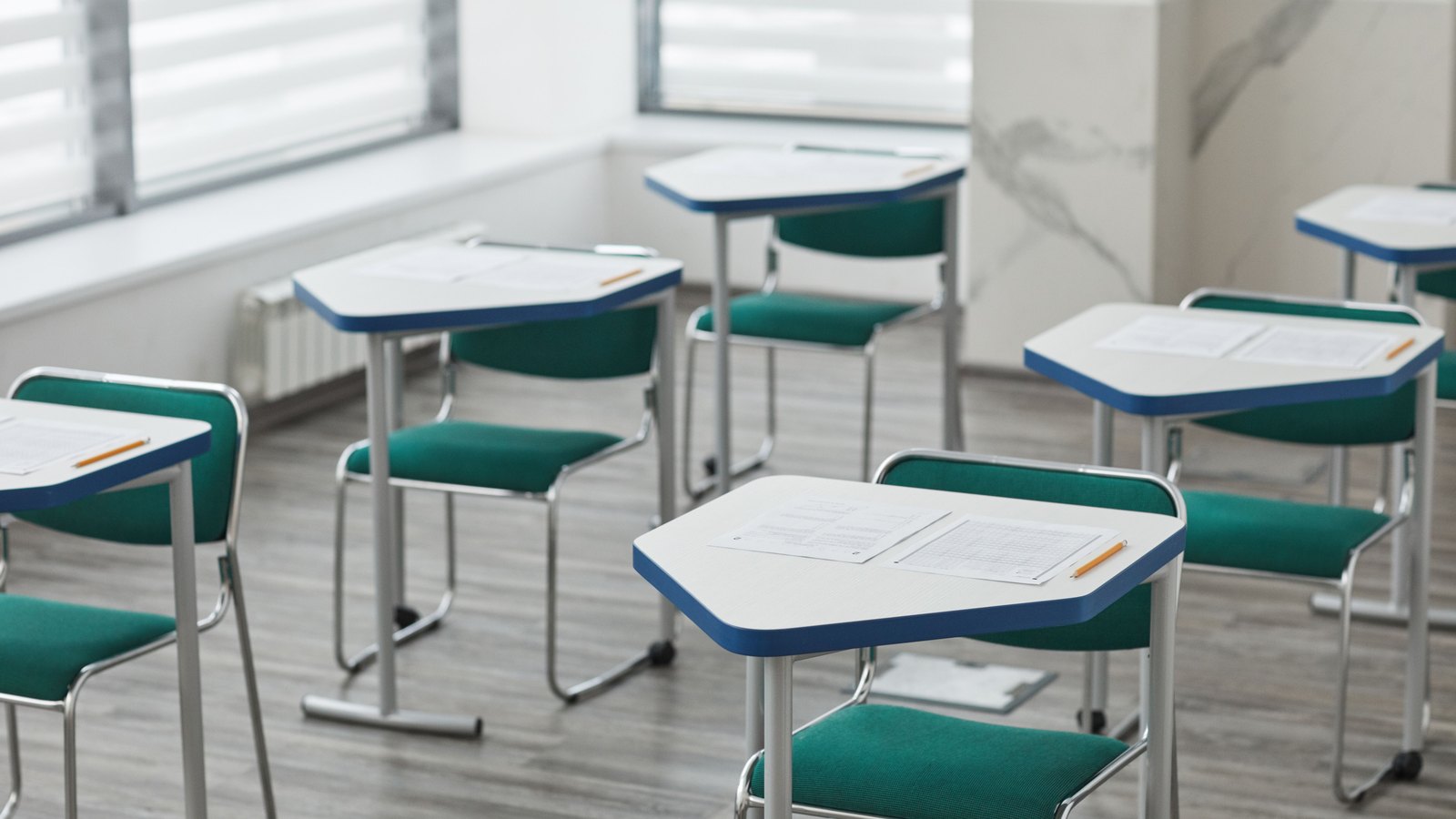 An empty classroom photographed at desk height. Half a dozen single desks with white tops, blue trim and green upholstered chairs stand in loose rows on a pale wood floor, each with a printed worksheet and a yellow pencil laid on it. Daylight comes through slatted blinds at the left. No person, no whiteboard writing, no signage, no school name and no legible text of any kind is in the frame. Illustration of an ordinary empty classroom, not a photograph of this event or this school.