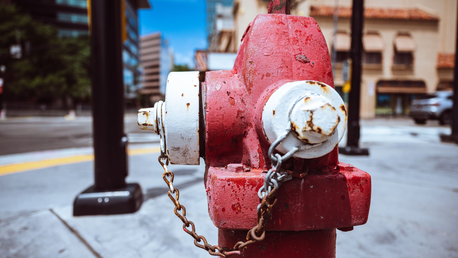 A close up of a weathered red and white fire hydrant on a pale concrete sidewalk, its rusted chain hanging from the capped side outlet, with a road, a kerb and a stucco building with a tiled roof and awnings blurred well out of focus behind it under a blue sky. No person, vehicle registration, house number, street sign, legible lettering, agency marking or identifiable address appears anywhere in the frame. A generic illustration of a street hydrant of the kind utility crews work on, not a photograph of any address under a notice.