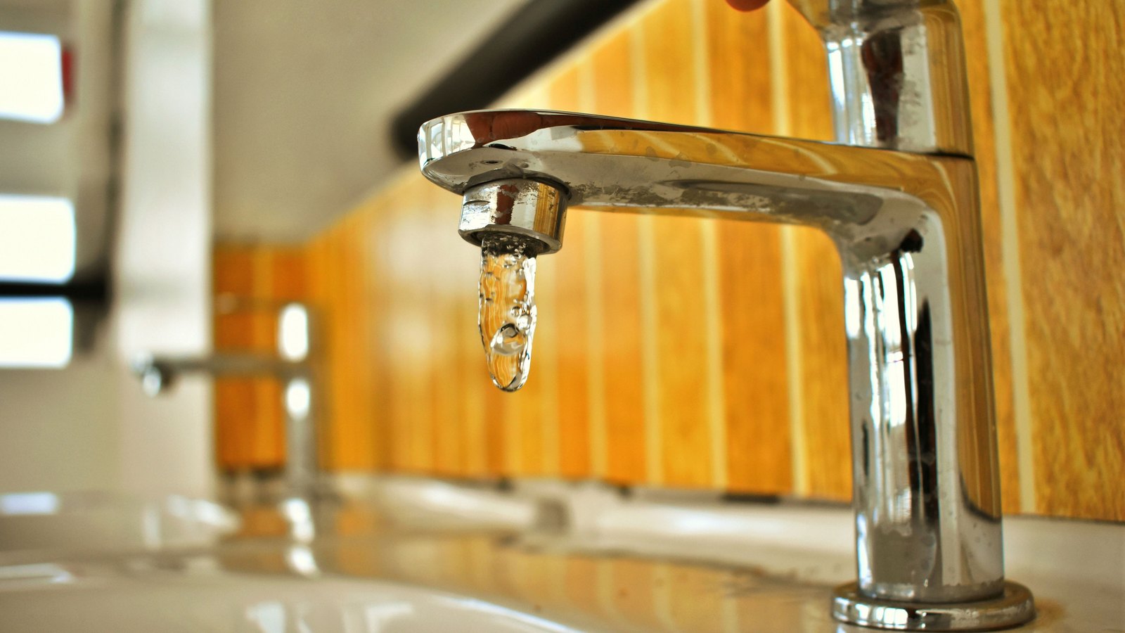 A colour close up of a chrome mixer tap with a single clear droplet of water hanging from its aerator, a white basin below it, a warm wood panelled wall behind it and a row of identical taps and basins receding out of focus to the left. A fingertip rests on the lever at the top edge of the frame. No faces, addresses, house numbers, plates, signs, branding or legible text of any kind appear anywhere in it. Stock photograph, not a photograph of this event.