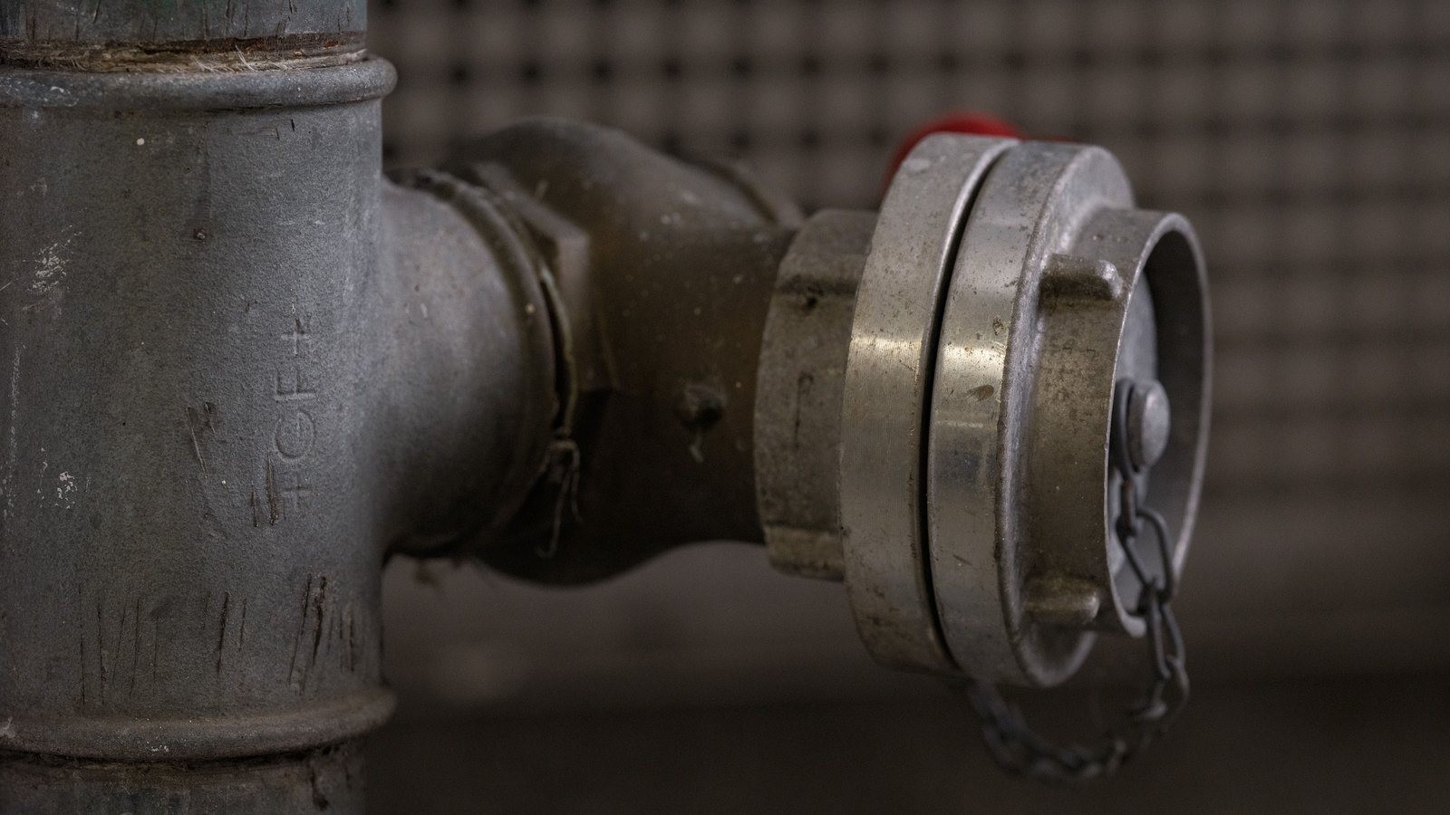 A close crop of a galvanised metal pipe tee joined to a capped coupling with a retaining chain, photographed against a dark blurred background. Faint raised casting marks are visible on the pipe body; there is no legible brand name, no signage, no person, no food and no identifiable place in the frame. A generic illustration of water pipework, not a photograph of Wittman Drive, of the broken main or of any address under notice, and not evidence about the condition of anyone's water. Its weakness is stated plainly: the fitting shown is an above ground coupling rather than the buried 4 inch main this item is about.
