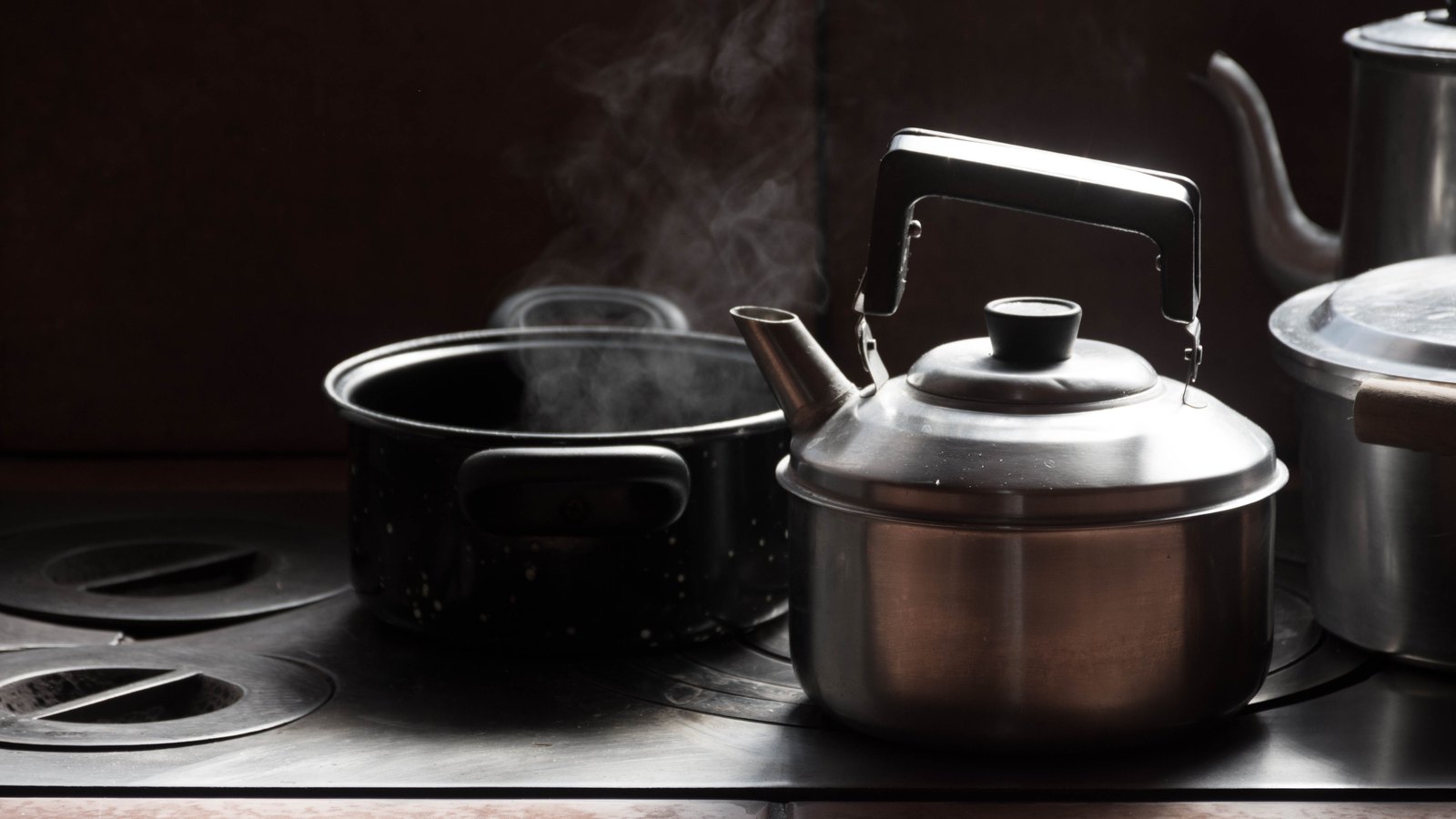 A stainless steel kettle stands on a dark stove top beside an open saucepan, steam rising from the pan into the shadow behind, with no people, signs or text in view.