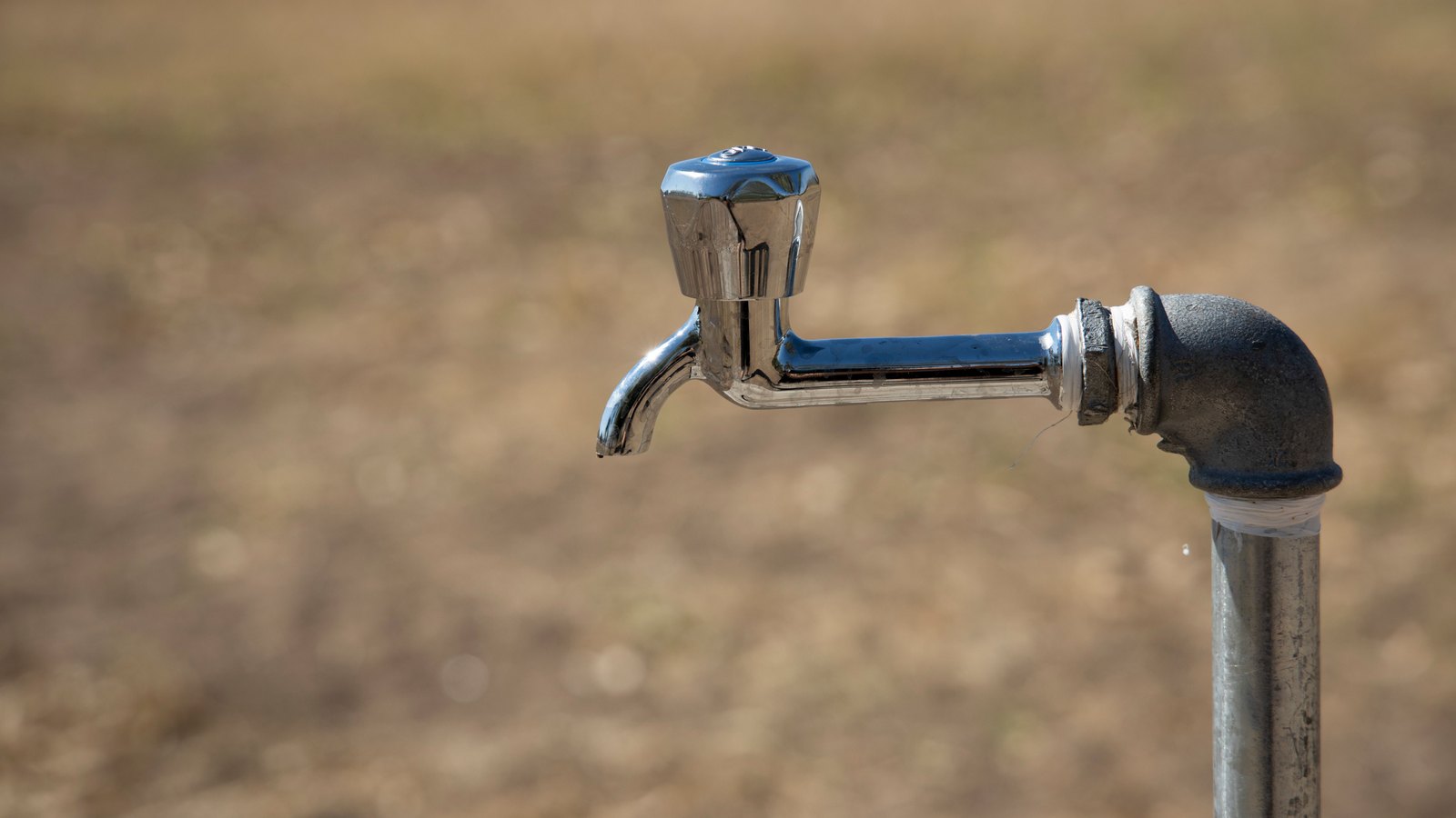A chrome outdoor tap with a fluted handle, mounted on a black elbow fitting at the top of a galvanised standpipe, against a blurred background of dry brown ground. No water is running from it. There is no text, signage, brand mark, person or identifiable place in the frame. A generic plumbing illustration, not a photograph of Lee County, Cape Coral or any address under notice, and not evidence about the condition of anyone's water.
