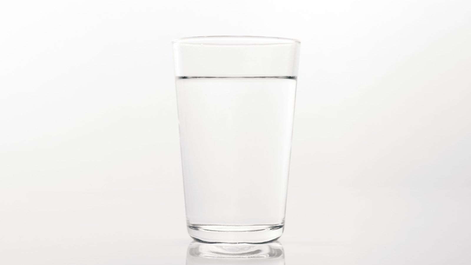 A single plain drinking glass, filled almost to the brim with clear water, standing on a white reflective surface against a plain white background. No people, buildings, taps, signage or lettering are visible and no place can be identified from it. A plain illustration of drinking water, not a photograph of any address or water system named in this item.