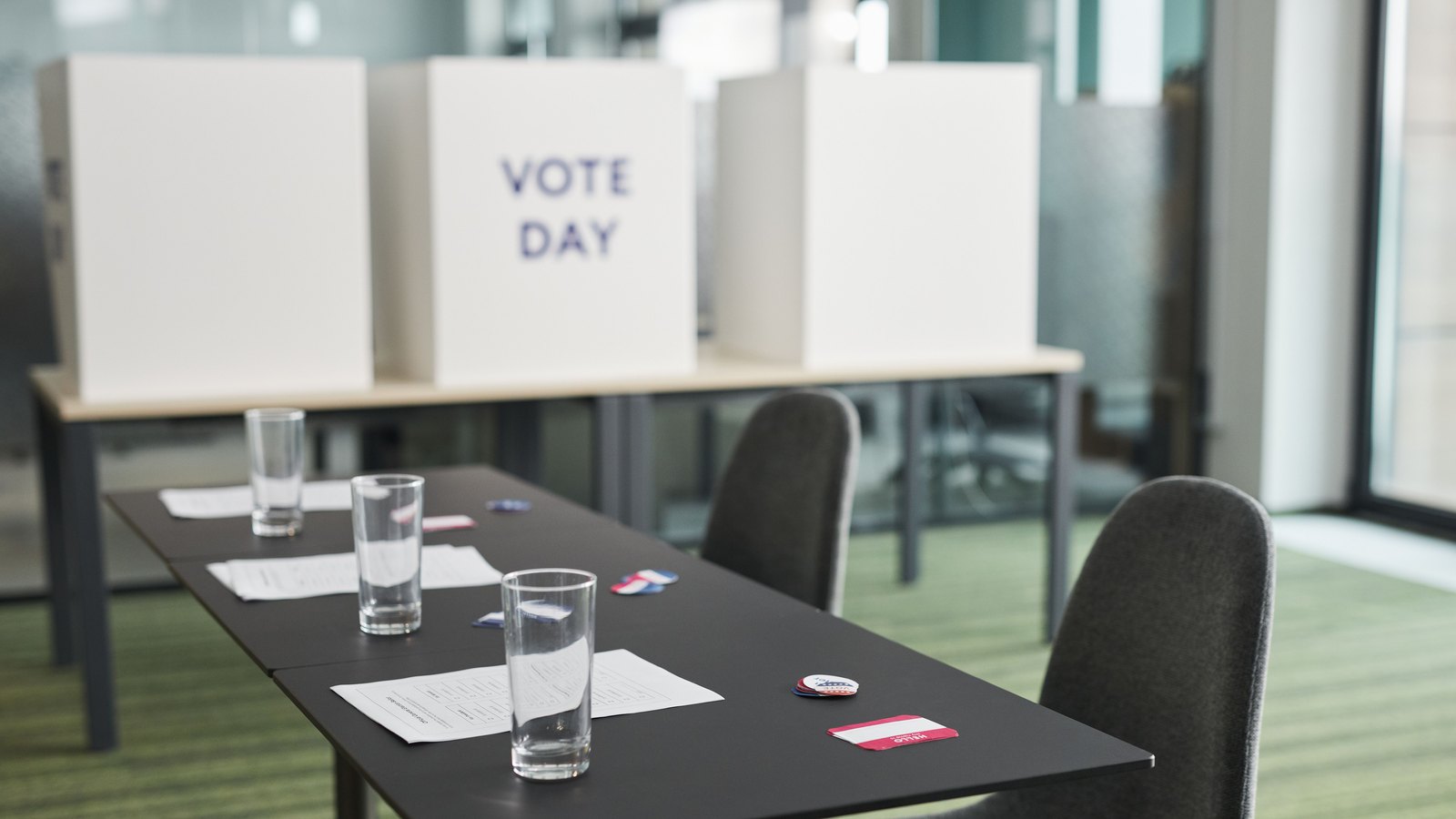 An empty polling room with white voting booth privacy panels reading VOTE DAY, and a table holding ballots, water glasses and I Voted stickers.