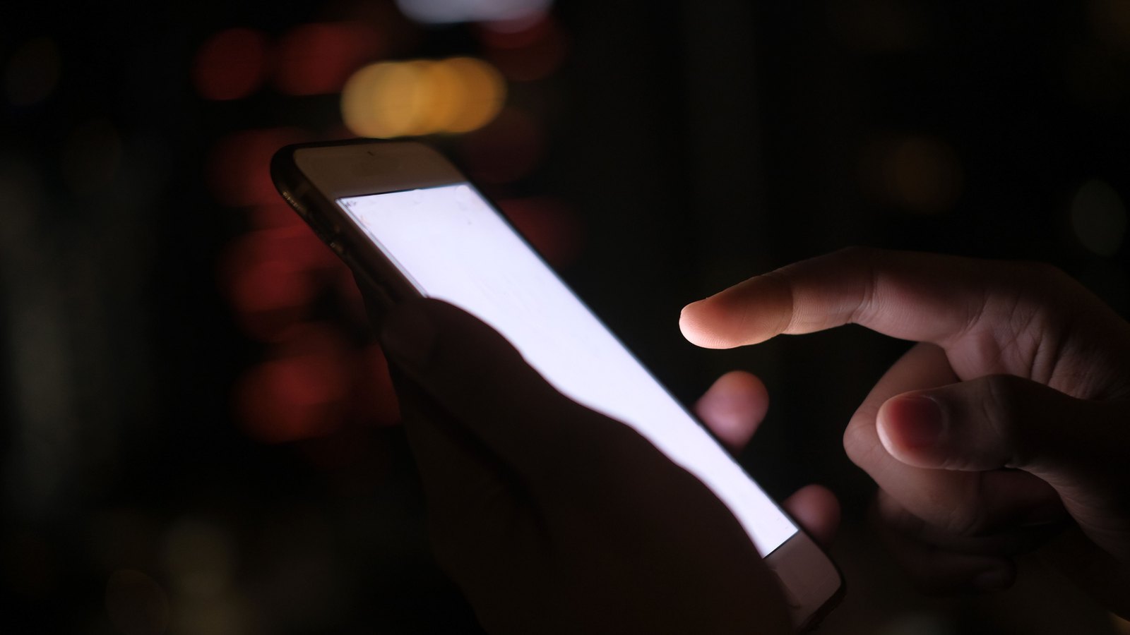 A pair of hands holding a smartphone in the dark, one finger poised over the screen, the display blown out to blank white so nothing on it can be read. The background is unlit bokeh. No face appears, and there is no app name, brand mark, logo, message text or legible text of any kind in the frame. A generic illustration of reading a message on a phone, not a photograph of this event.