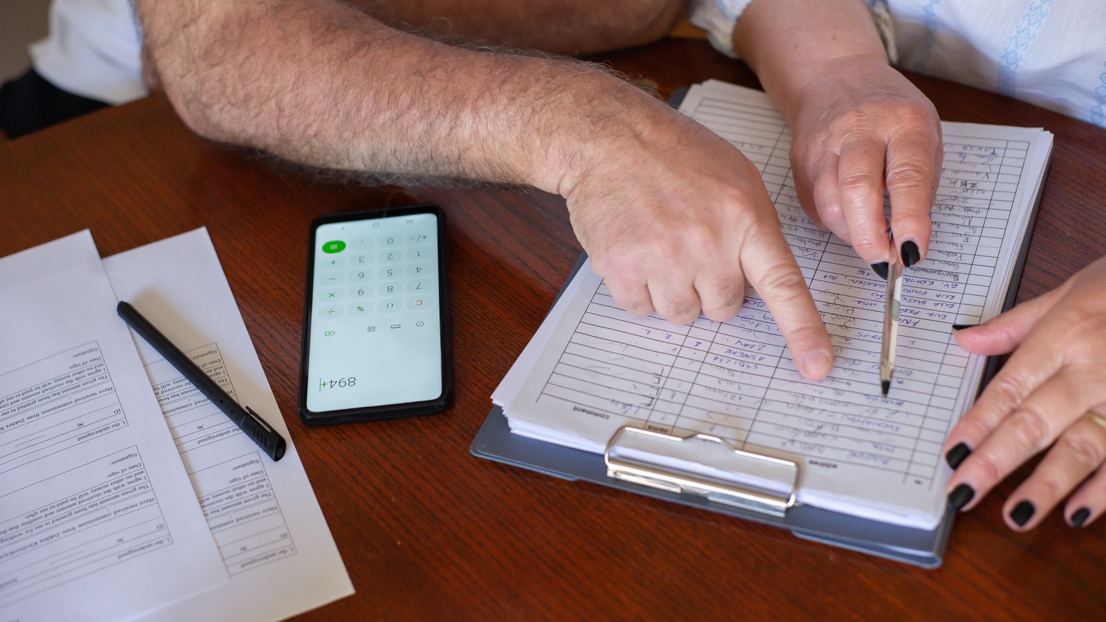 Two pairs of hands at a wooden table working through paperwork, one finger tracing a line on a clipboard while the other hand holds a pencil, a phone calculator and a pen beside a printed form. No face appears in the frame, and no letterhead, logo, name, account number or legible text of any kind can be read on any of the documents. Stock photograph, not a photograph of this event.