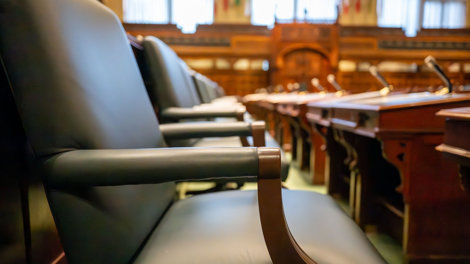 Empty dark leather armchairs along a row of wooden desks fitted with slim microphones inside a wood panelled legislative chamber, the far end of the room thrown out of focus. No person is in frame and no lettering, seal or sign is legible. Stock photo, not a photograph of this event.