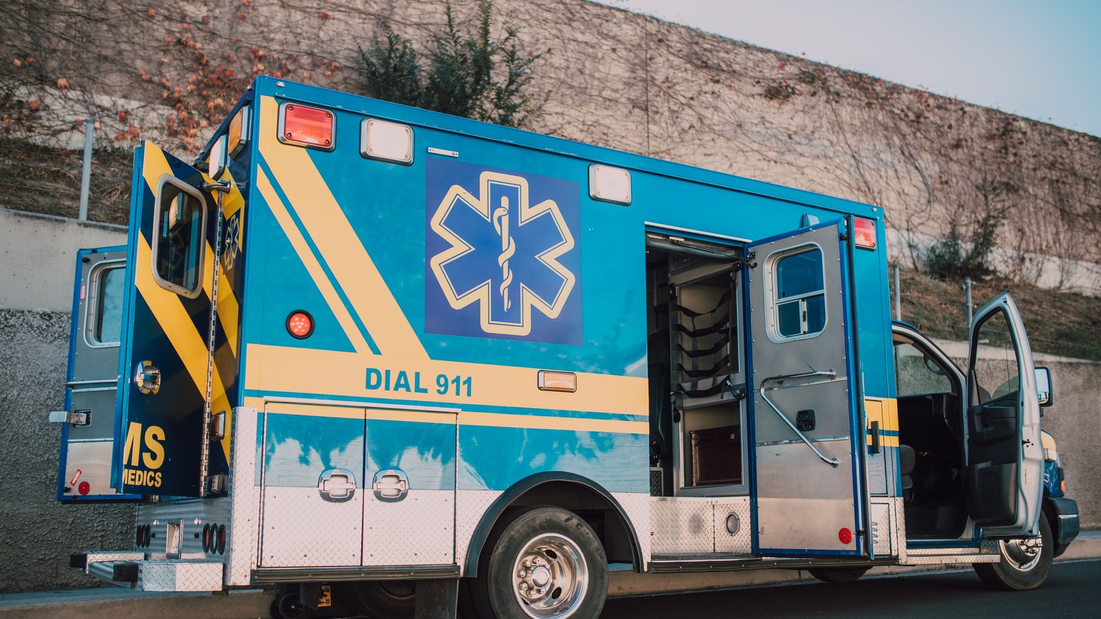 The side of an American ambulance parked beside a plain concrete retaining wall in low afternoon light, its side compartment door standing open on empty equipment shelving, a blue Star of Life on the body panel above the words DIAL 911, and generic medic lettering on the rear quarter. No person, number plate, street name, address or agency name appears anywhere in the frame. A generic illustration of an ambulance of the kind sent to a crash with injuries, not a photograph of this event.