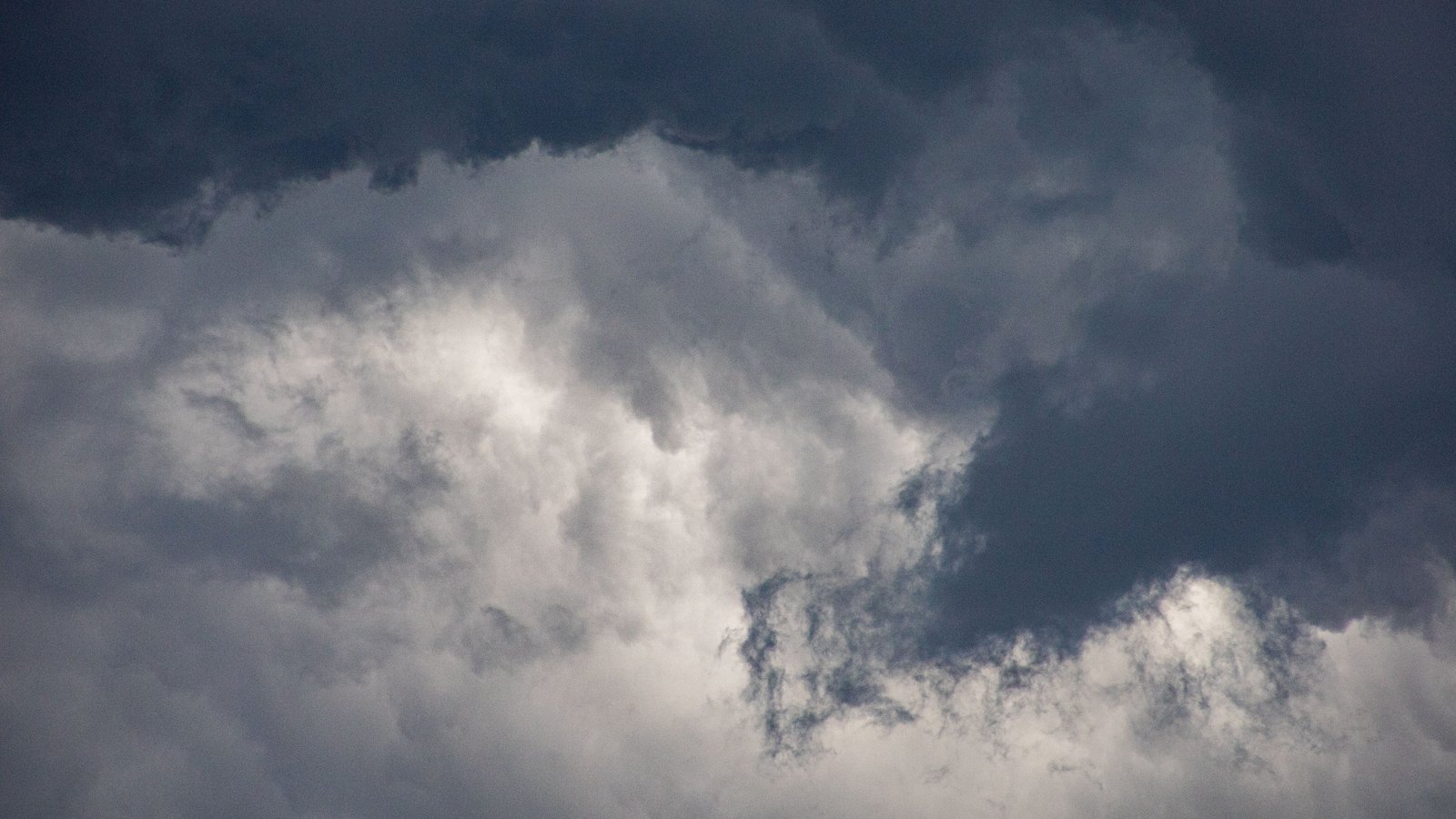 A bank of dark grey storm cloud filling the frame, with a paler cloud mass lit from behind and no horizon, buildings or text in view.