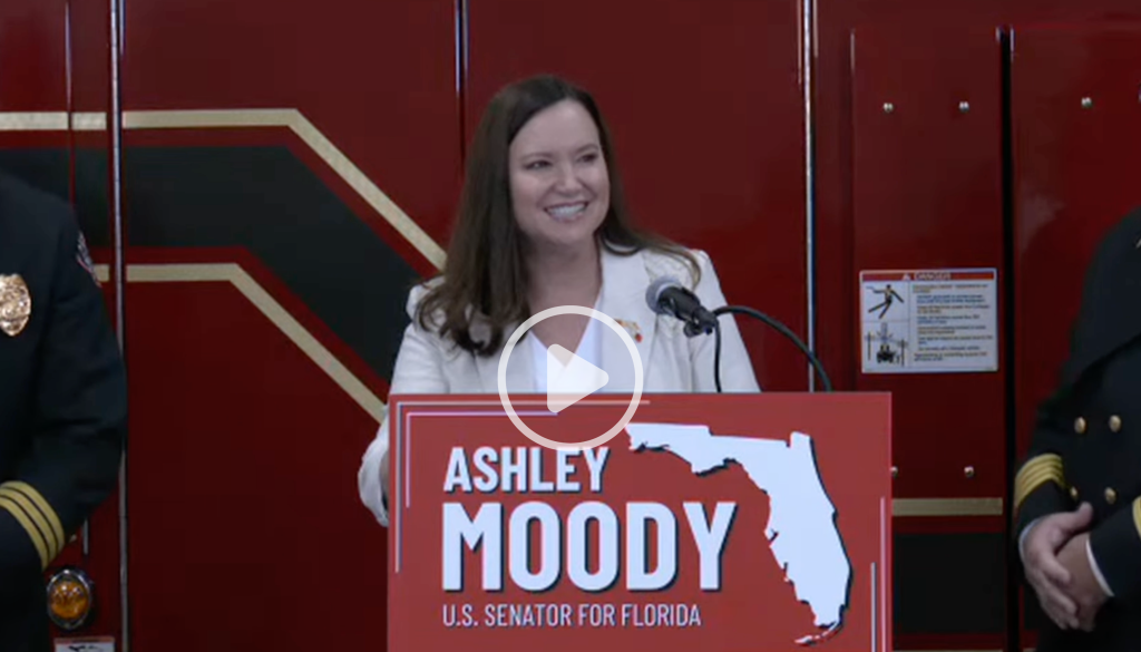 Senator Ashley Moody speaks at a lectern in front of a red fire engine during the Lehigh Acres grant announcement.