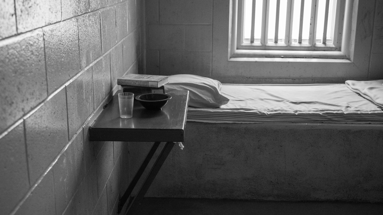 A bare cell interior in black and white: a concrete bunk with a thin mattress and folded blanket, a small steel shelf holding a plastic cup, a bowl and two books, a breeze-block wall and a narrow barred window throwing flat daylight across the floor. No person, no officer, no vehicle, no building exterior, no address, no signage, no agency name and no other identifying mark appears in the frame. A stock photograph, not a photograph of this event.