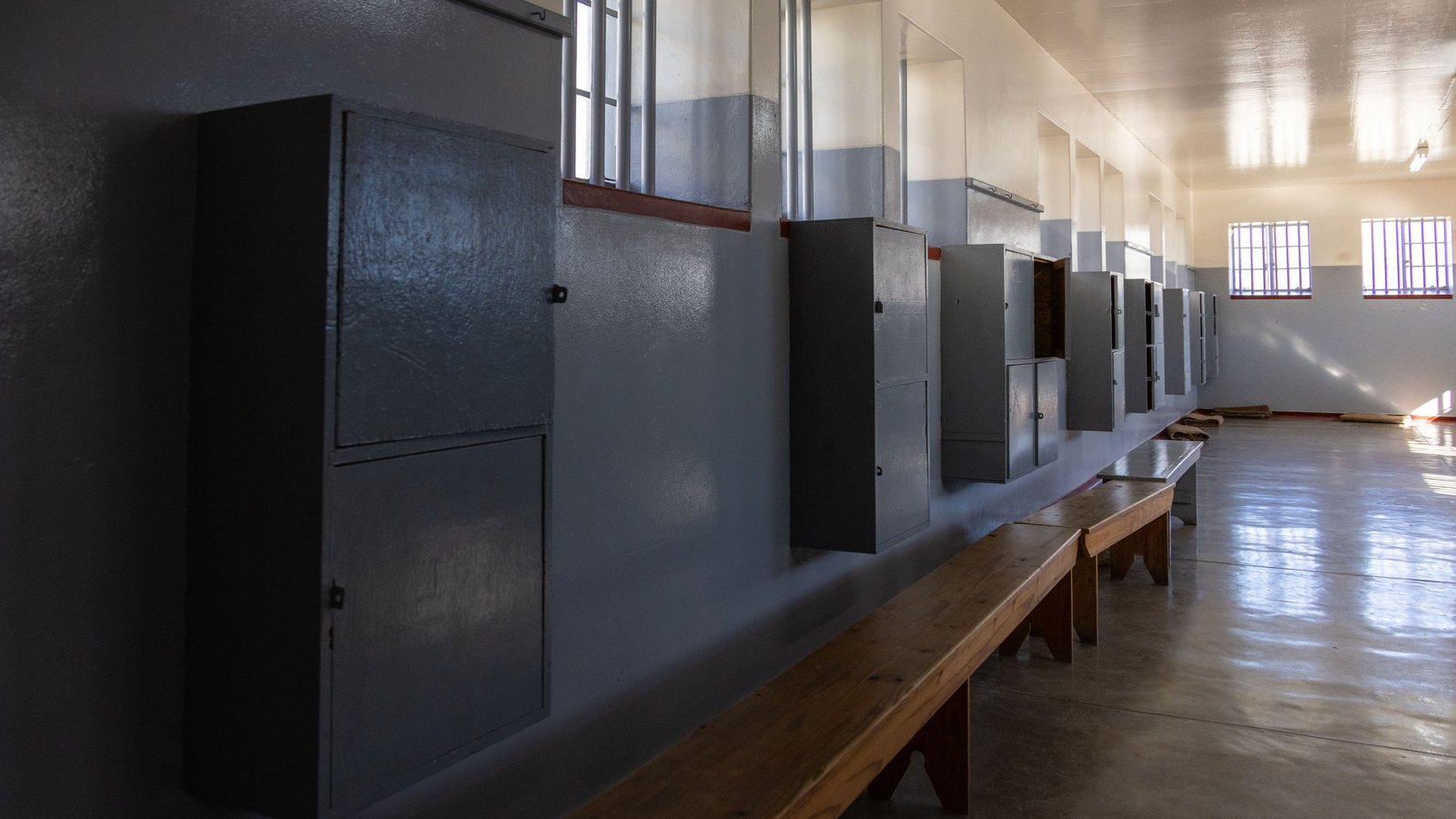 A long empty corridor inside a detention block, with a plain grey painted wall, a row of small dark metal lockers fixed above a low wooden bench that runs the length of the room, barred windows high on the wall throwing daylight across a bare concrete floor, and no people, signs, text, insignia or brand marks legible anywhere. A generic illustration, not a photograph of this event.