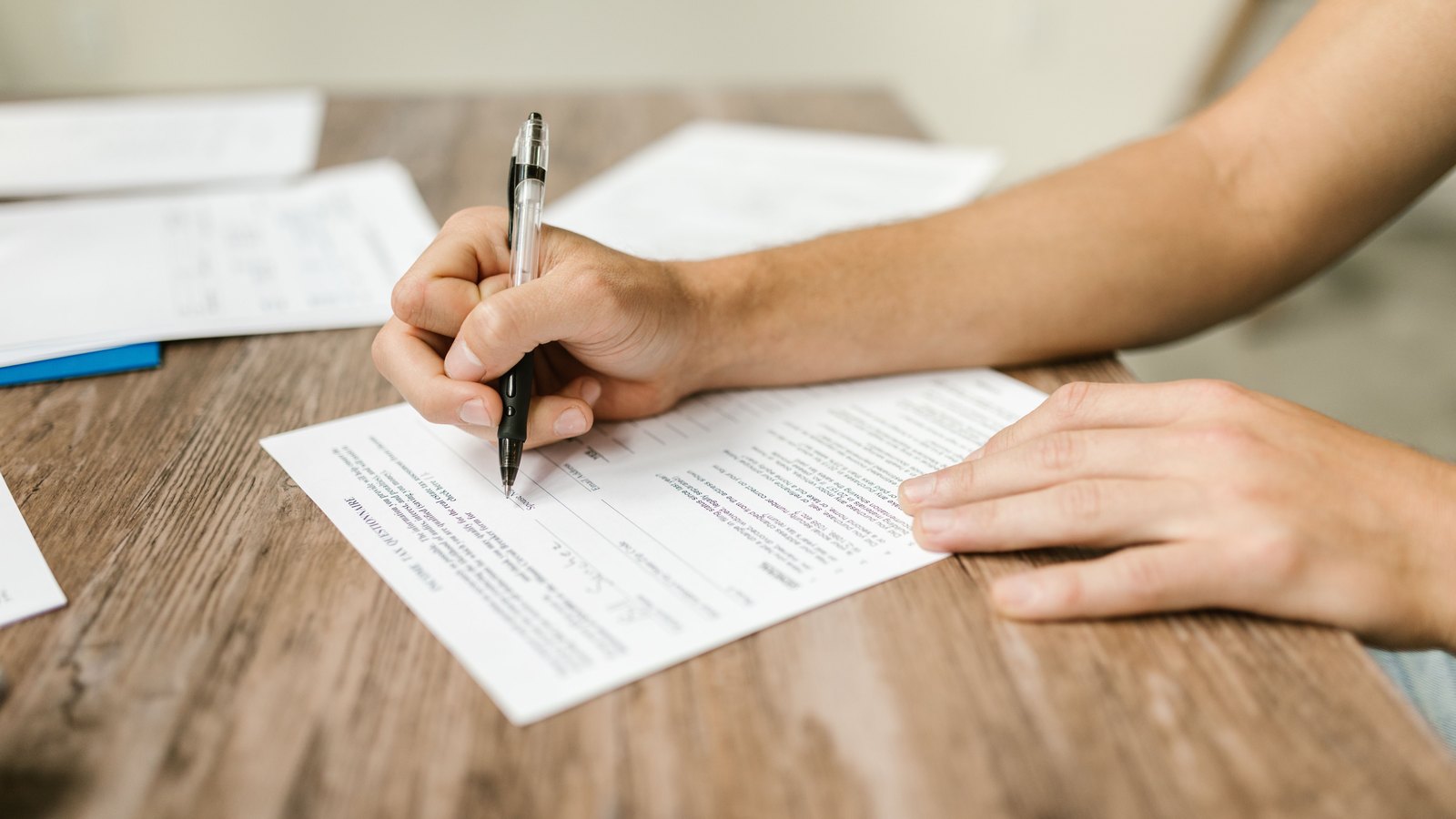 Hands fill out a paper form with a ballpoint pen on a wooden table, with no face, readable text, sign or location visible. A generic paperwork scene, not a photograph of this event.
