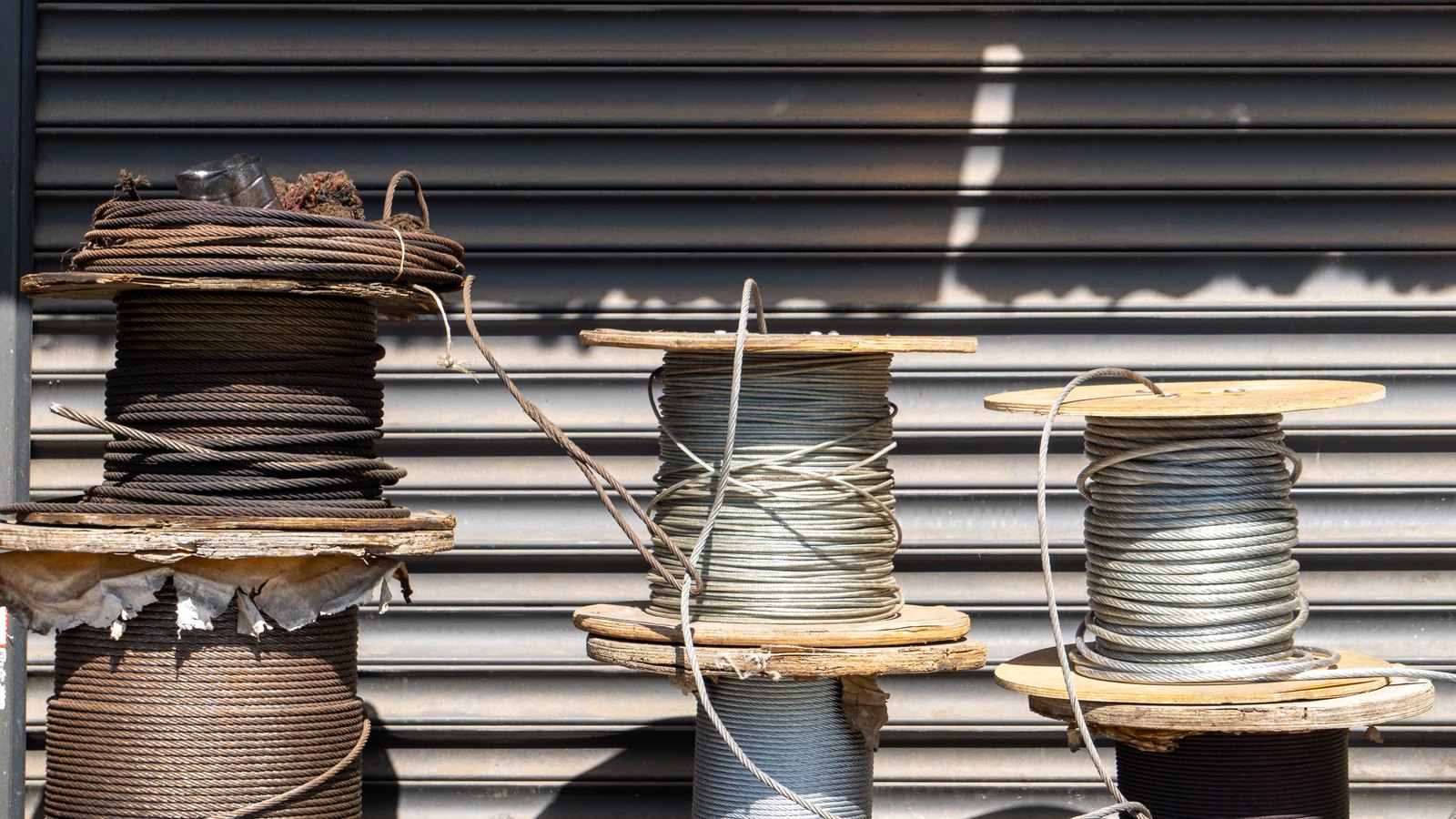 Four large wooden-flanged spools of stranded metal cable stand in a row on a sunlit pavement against a corrugated grey roller shutter. The spool on the left is wound with dark rust-brown wire rope and its wooden flange is split and weathered; the two to the right carry brighter galvanised strand, with loose loops of cable hanging free and casting hard shadows on the metal behind. No people, signage, text or brand marks are in view. A generic illustration of cable stock, not a photograph of the damaged fibre optic lines in Lehigh Acres.