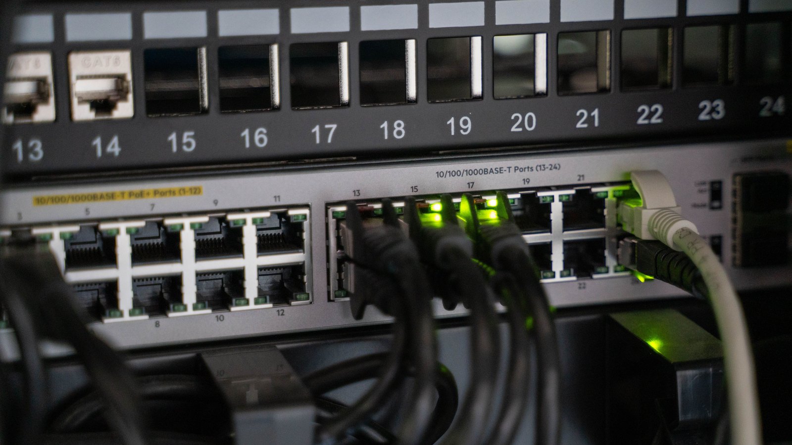 A network patch panel above a switch in a dim rack, ports numbered 13 to 24 across the top, with grey and black ethernet cables plugged into the lower row and several link lights glowing green.