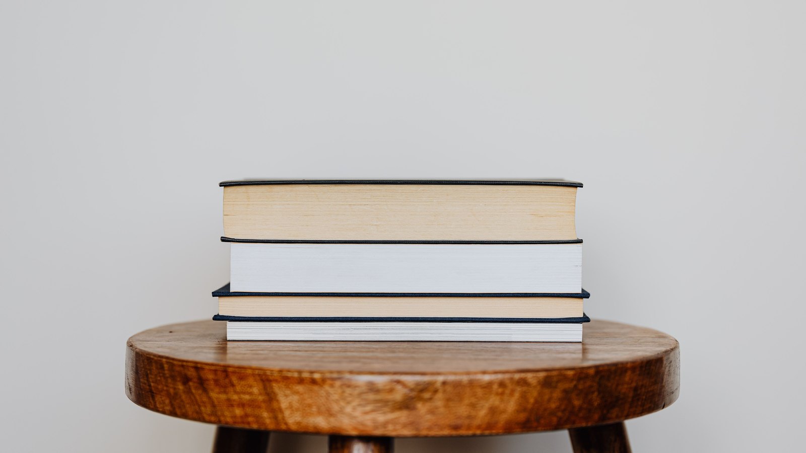 Three closed hardcover books stacked on a round wooden stool against a plain pale grey wall. No people, text, signage, brand mark or identifiable place is visible, and the books carry no readable titles. A neutral illustration, not a photograph of this case, this courthouse or any person connected with it.